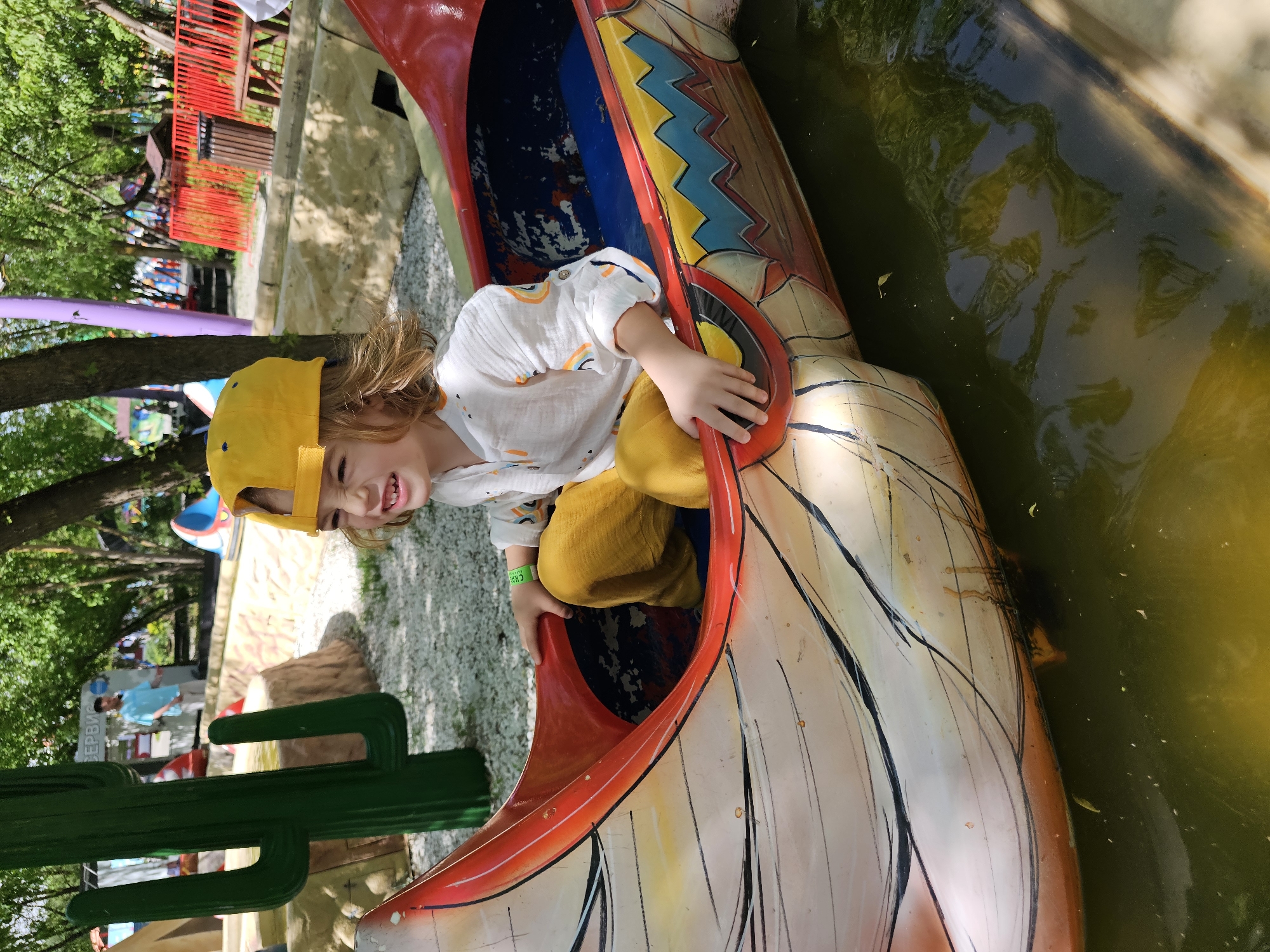 A child wearing a yellow cap and yellow pants sits inside a boat-shaped playground equipment. The equipment is decorated with various colors and designs. The boat appears to be floating in a body of water. A large, wooden cactus prop is nearby. The location is Khimki, Russia.