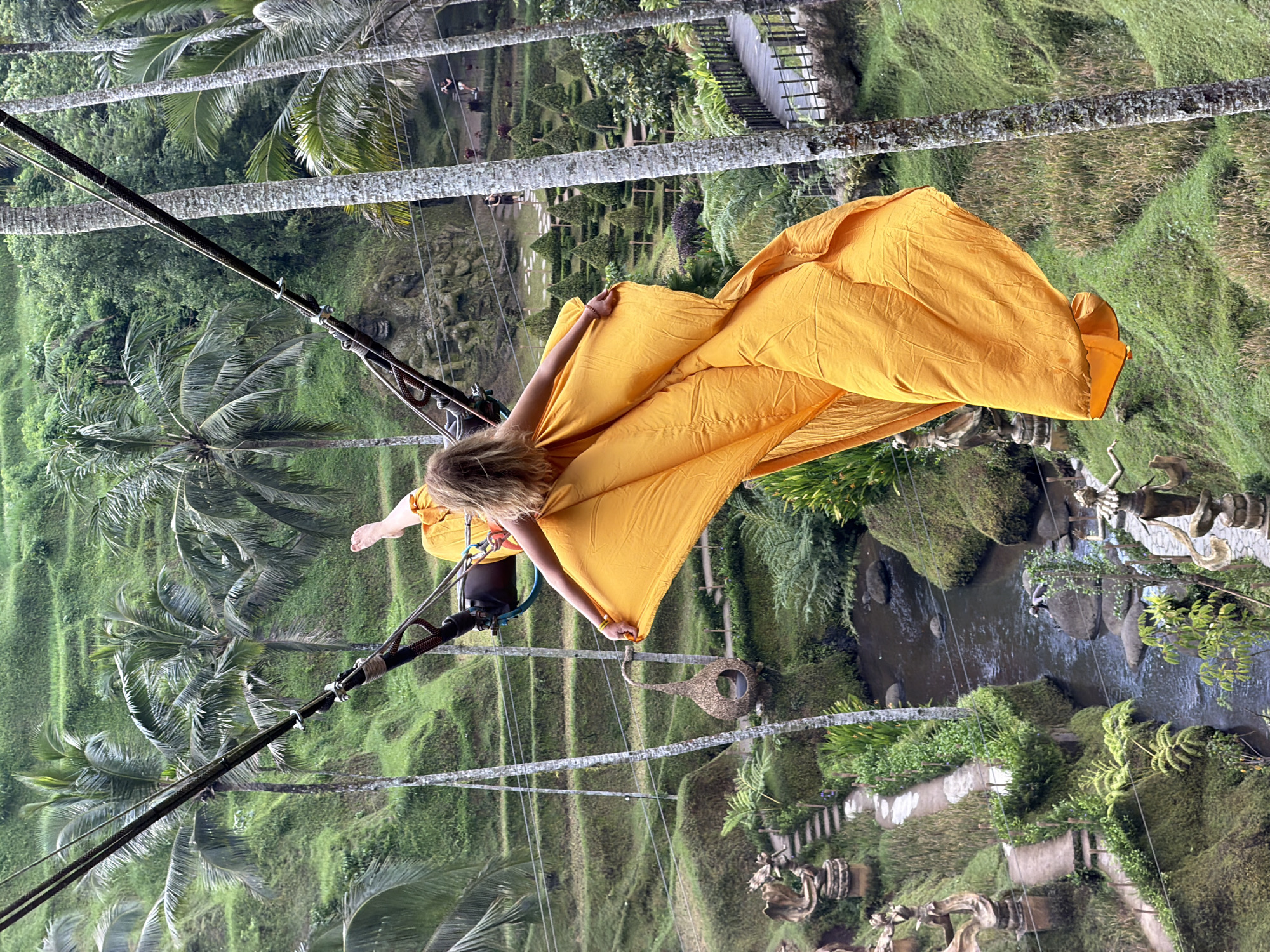 A woman swings over a green landscape in Tegal Suci, Indonesia. The woman is suspended by a metal contraption that hangs from two thick ropes. She wears a safety harness and holds a large piece of golden-yellow fabric that billows around her. The landscape includes rice paddies or terraces, palm trees, and a small stream. There are also pathways, sculptures, and a wooden bridge visible in the background. The image appears to be taken from a high vantage point, looking down at the scene.