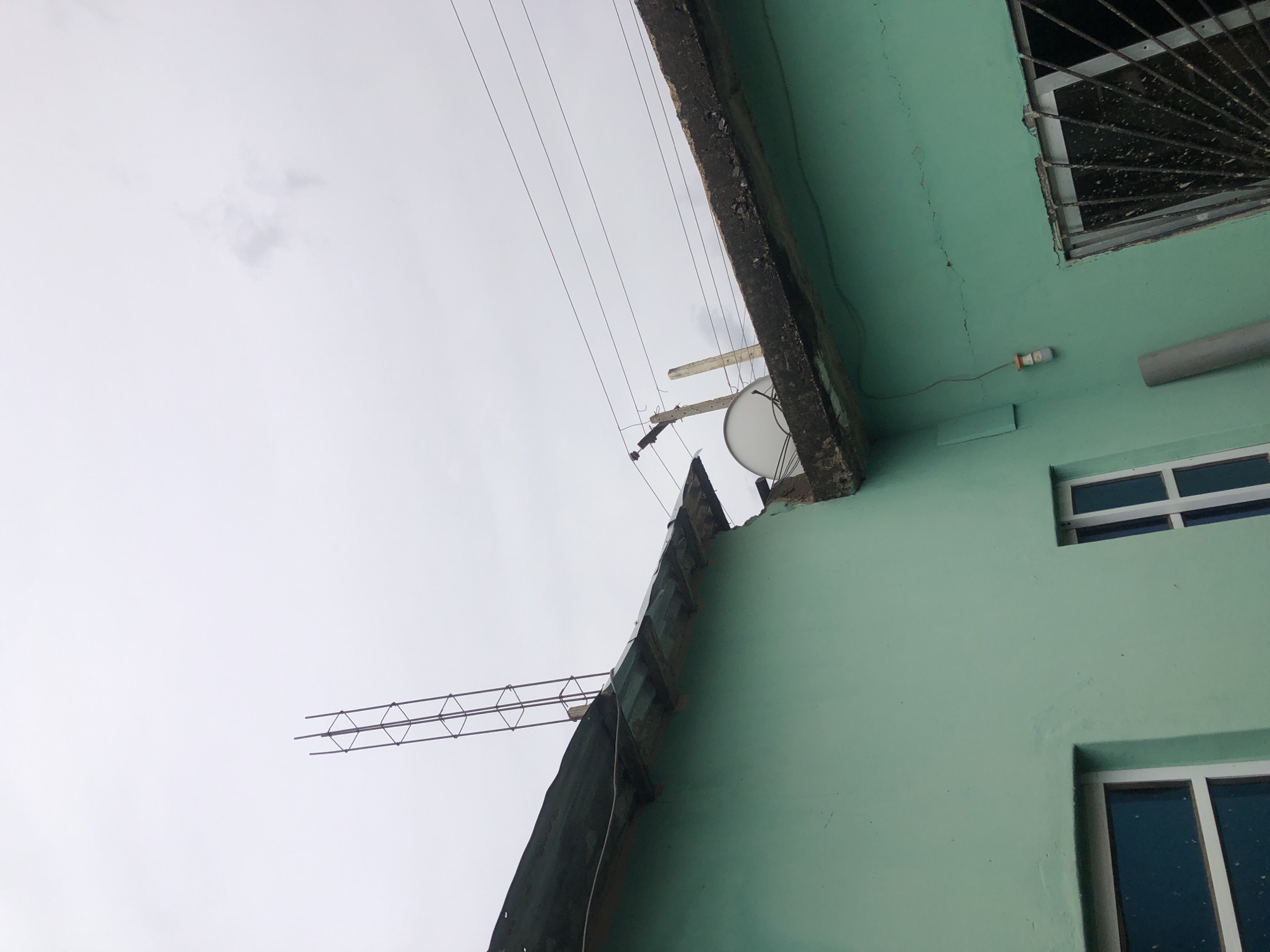 The image presents an upward view of a light green-painted building facade against an overcast, bright sky. The building appears tilted due to the camera's orientation.
The primary subject is the building's exterior, characterized by its faded, cracked, light green walls. Several windows are visible; one in the upper right corner features dark security bars, appearing dirty or corroded. Below it, a clean, modern-looking window with white frames and dark glass is present, and another similar window is partially visible towards the bottom right.
A rough concrete ledge or roofline runs diagonally across the image, separating the building from the sky. Mounted near this ledge is a small, white satellite dish. Above the building, multiple utility wires stretch horizontally across the frame, supported by what appear to be wooden poles, one of which is clearly visible. Further to the left, a section of metal rebar, forming a triangular lattice structure, extends into the sky, suggesting ongoing construction or an unfinished element.
Attached to the green wall is a small, cylindrical light fixture connected by a wire, and a grey pipe runs horizontally below the security-barred window. The sky is uniformly light grey, indicating an overcast day with diffused lighting, and faint cloud formations can be observed. There are no people or discernible activities taking place, and no visible text. The overall impression is one of an older, perhaps residential, building in an urban or semi-urban setting, captured from a unique, rotated perspective.