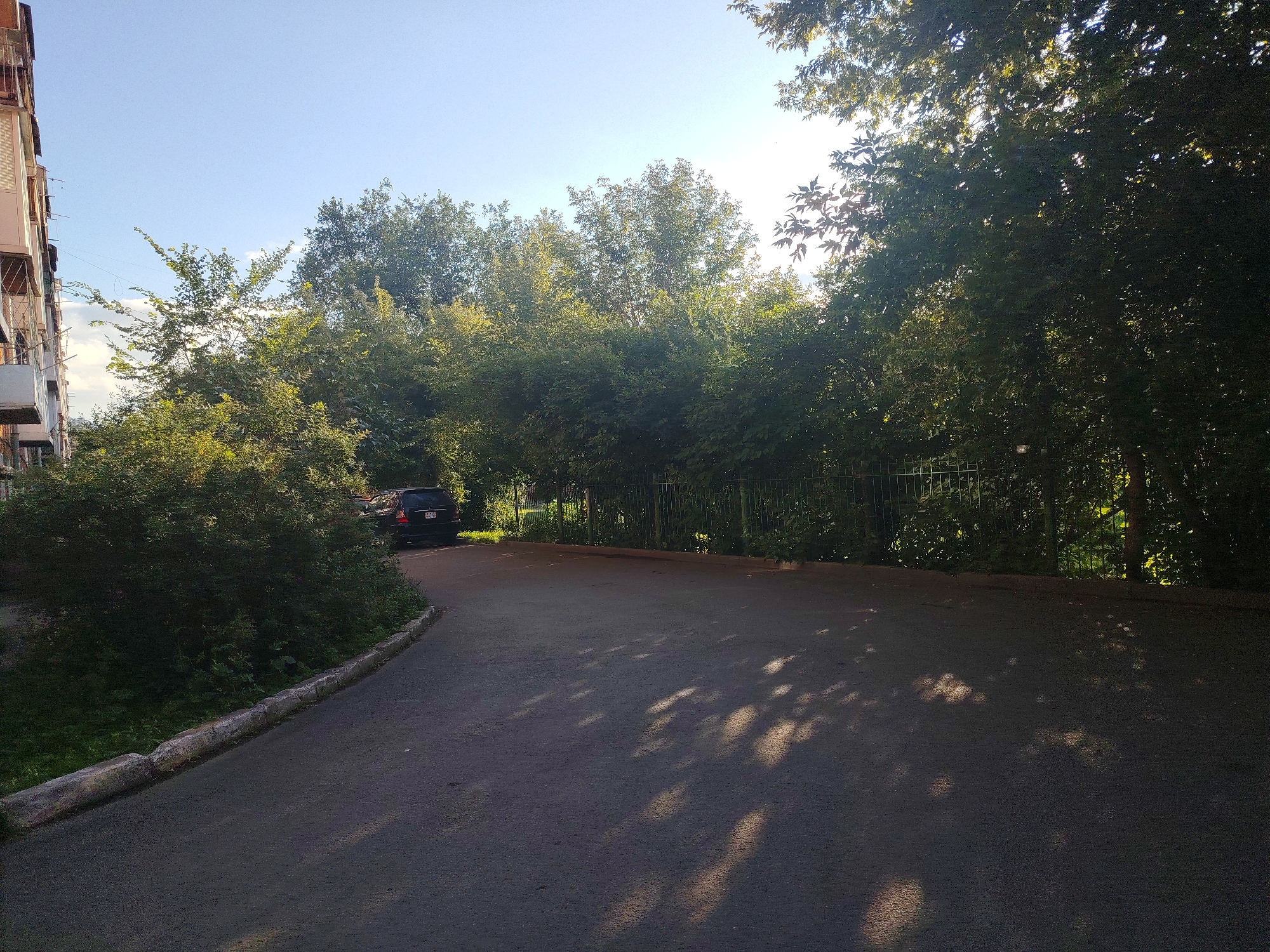 The image depicts a residential courtyard in the city of Kemerovo, Russia. The scene is dominated by a paved road curving through the frame, bordered by lush green trees and bushes. A dark-colored SUV is parked on the right side of the road, partially obscured by foliage. In the background, a chain-link fence runs along the edge of the property, separating it from more trees and vegetation. To the left, a multi-story building with balconies is visible, its facade partially illuminated by sunlight. The sky is a clear blue, with dappled sunlight filtering through the leaves of the trees, casting shadows on the road. The overall impression is one of a peaceful, sunlit afternoon in a typical urban residential area.