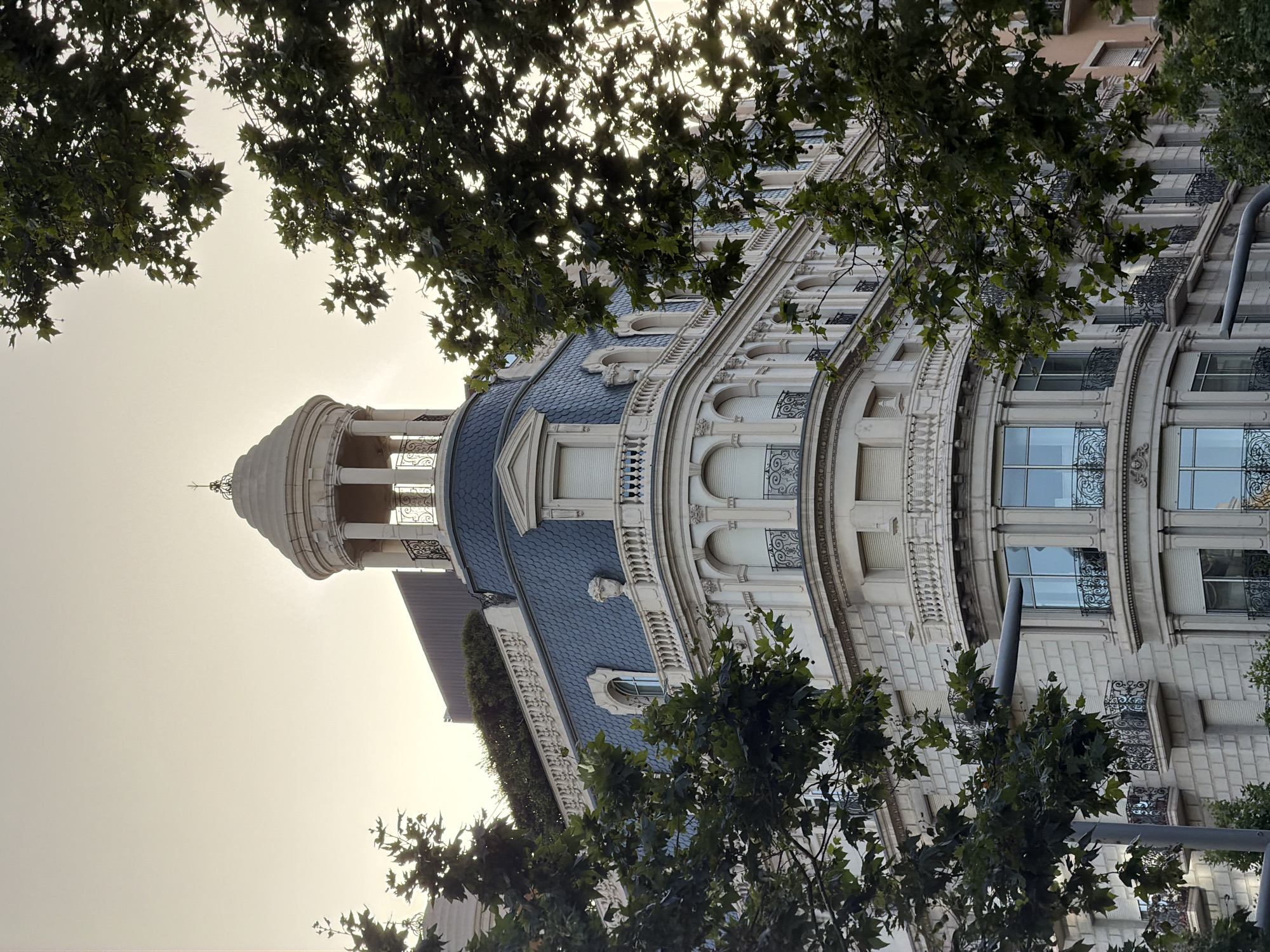 This is a photograph of a historic building in Barcelona, Spain, with trees in the foreground. The building is tall and ornate, with a distinctive mansard roof covered in dark blue tiles. A domed cupola with columns crowns the top of the building. The facade is made of light-colored stone and features multiple balconies with decorative iron railings and large windows. The sky is a pale, hazy yellow, suggesting it may be dawn or dusk. The trees, with their green leaves, partially obscure the view of the building. The overall impression is one of architectural grandeur set against a natural backdrop within an urban environment.