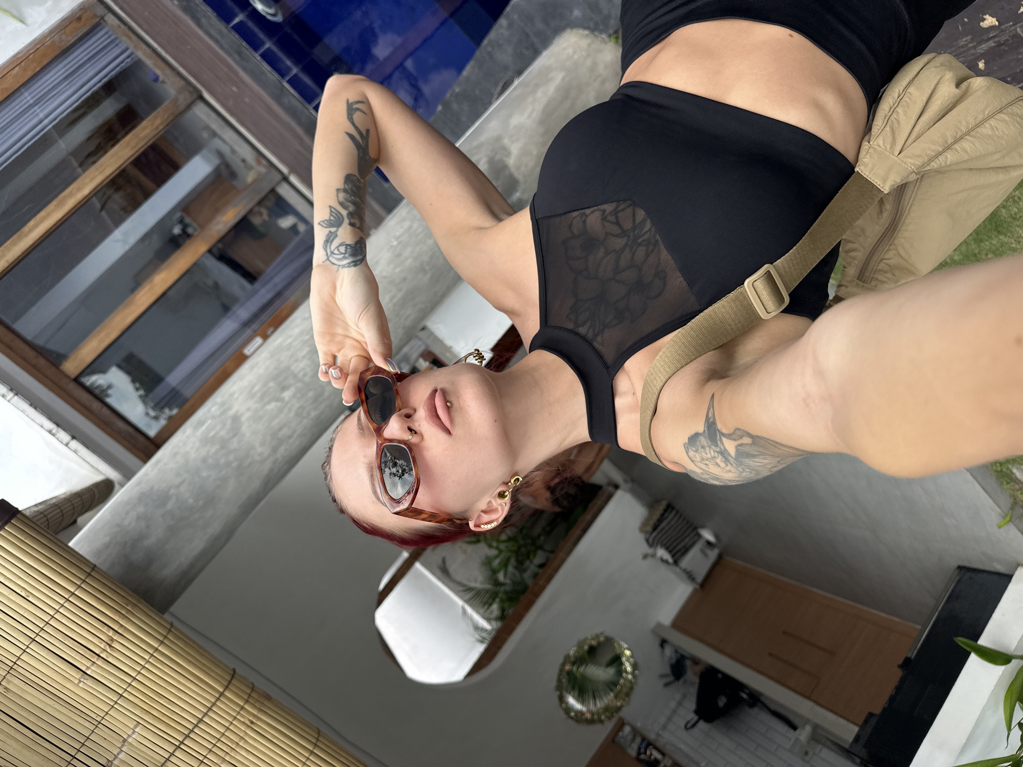A medium shot captures a woman, upside down, wearing a black sports bra with sheer mesh panels, black shorts, and sunglasses, along with a beige shoulder bag. The woman has fair skin, red-tinted hair, a nose ring, a lip piercing, and multiple earrings. She has tattoos on both arms. She is holding her sunglasses.
Behind her, there are various elements, including a window, bamboo, and an architectural structure, suggesting an outdoor or semi-outdoor setting. The location is in North Kuta, Indonesia.