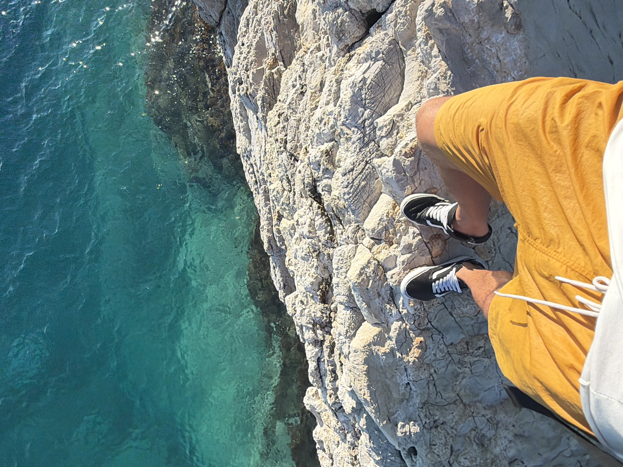 The image shows a person's legs and feet from a high-angle perspective, looking down towards the sea. The person is wearing yellow shorts and black and white sneakers, and is standing on a rocky outcrop overlooking clear, turquoise water. The rocks are light-colored and textured, with some dark patches where the water meets them. Sunlight glints off the surface of the water, creating small sparkles. The background is dominated by the vast expanse of the sea, with a darker, rocky underwater area visible beneath the surface. The scene is likely located in Petrovac centar, Montenegro, given the characteristic coastal landscape.
