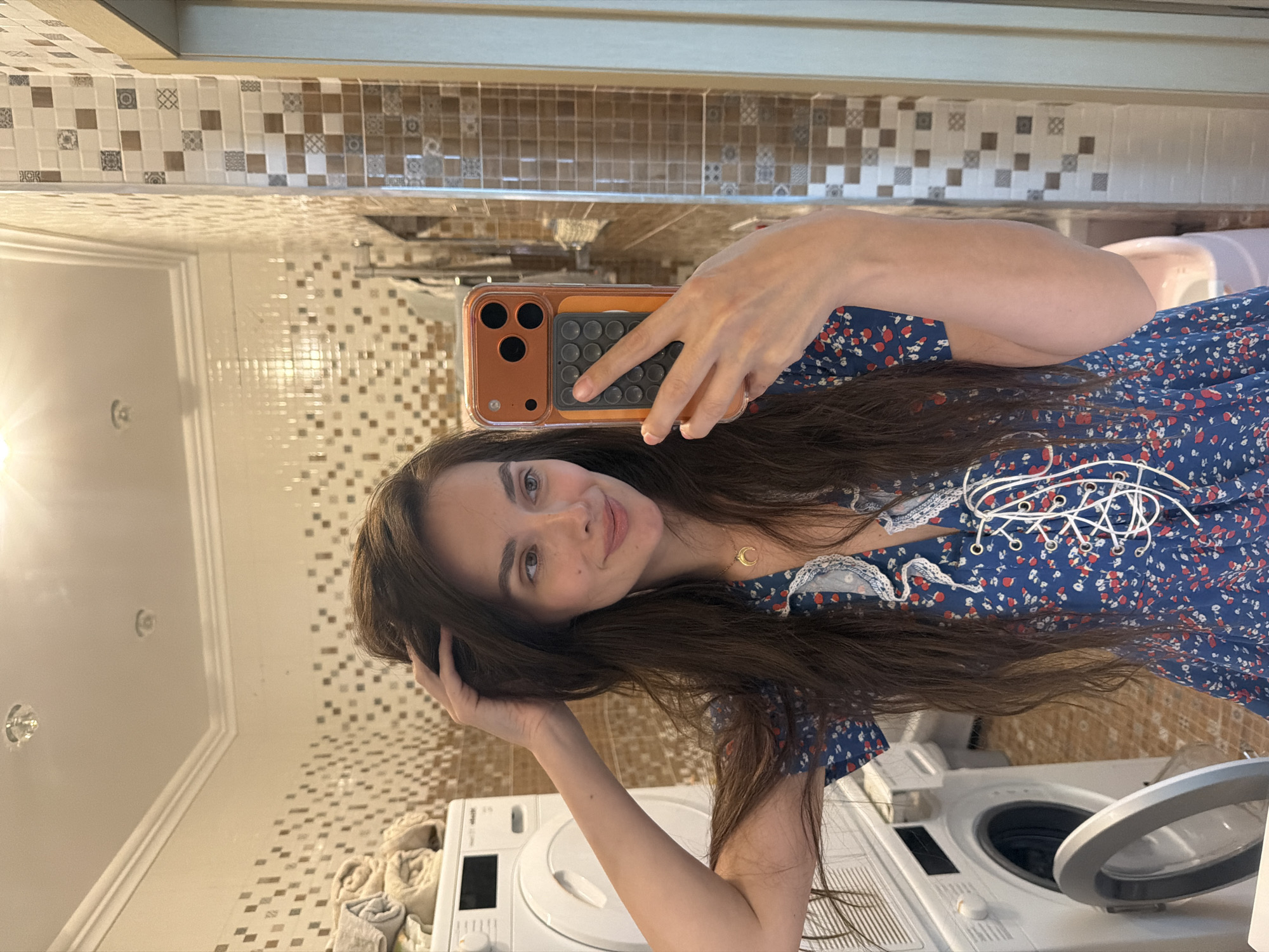 A woman takes a selfie in a bathroom. She has long, dark brown hair and is smiling. She is wearing a blue dress with a red and white floral pattern. She is holding her phone in her right hand, and her left hand is touching her hair. In the background, there is a tiled wall, a mirror, a washing machine, and a dryer. The overall impression is one of a casual, everyday moment. The tiling in the bathroom suggests a modern or updated style. The presence of laundry appliances indicates this is likely a residential bathroom, possibly in a home or apartment. The lighting in the image appears to be artificial, suggesting it could be any time of day. The woman's expression is pleasant and relaxed. The location context is Ufa, Russia. No visible text is present in the image.