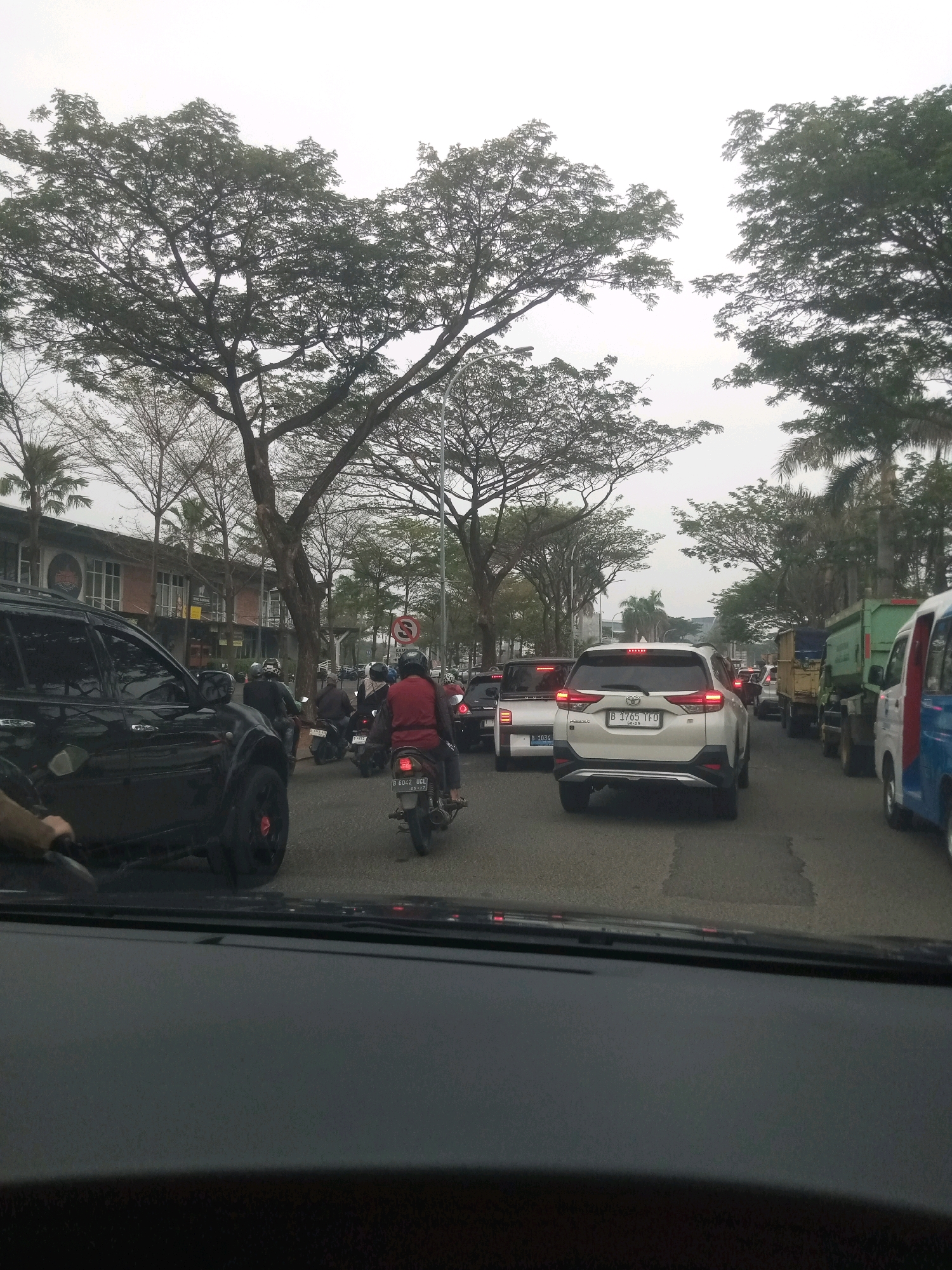 This is a photograph taken from inside a vehicle in moderate traffic on a street in East Jakarta, Indonesia. The scene depicts a road lined with trees, some of which are bare of leaves. On the left side of the road, there are buildings with shops or businesses. The street is congested with vehicles, including cars, motorcycles, and trucks.
In the foreground, the dashboard and windshield of the vehicle from which the photo is taken are visible. A black SUV is prominent in the left lane. Moving further down the street, several motorcycles are seen navigating through the traffic. One motorcycle rider in a red jacket is particularly noticeable. In the right lane, a white SUV with the license plate B 1765 TFO is stopped. Beyond that, there are several trucks, including a green one, and a blue and white van. Traffic lights and street signs are present, including a red circular sign with a diagonal line indicating no right turn. The sky is overcast. The overall impression is a typical urban traffic scene in Jakarta.