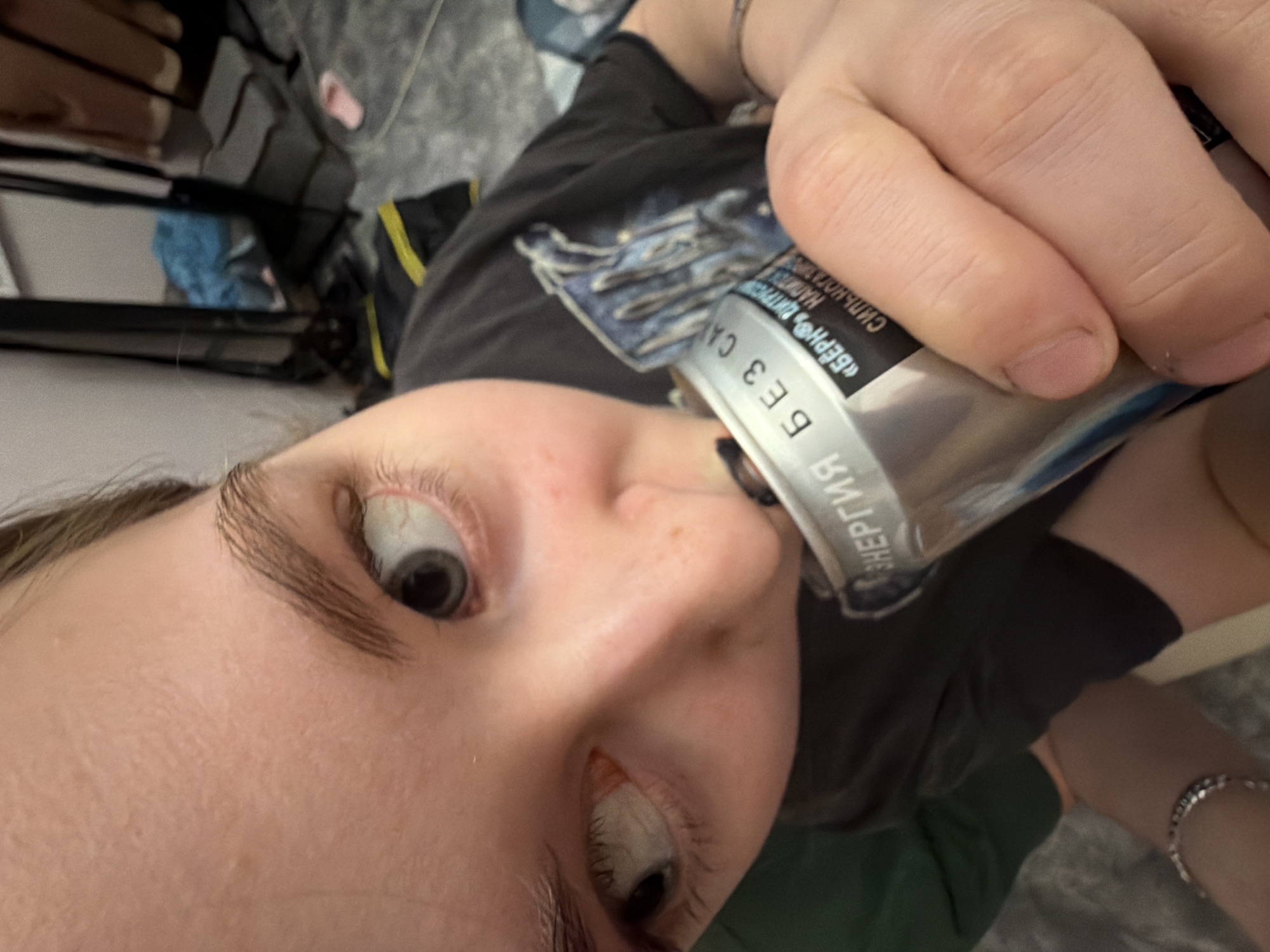 A low-angle, close-up shot captures a person drinking from a can in what appears to be an indoor setting. The person, positioned in the foreground, looks directly down towards the camera with wide-open eyes, revealing dark pupils and visible sclera. Their face shows natural skin texture, and prominent, well-defined eyebrows.
They are holding a silver and blue can to their mouth with one hand. The can features Russian text, including "БЕРН® ЭНЕРГИЯ" (Bern® Energy) and "БЕЗ САХАРА" (Without Sugar), indicating it is likely an energy drink. The person wears a dark t-shirt, part of which has a graphic design that is partially obscured by their hand and the can. A silver bracelet is visible on their wrist.
In the background, out of focus, are dark shapes that suggest furniture or shelving, and a glimpse of blue fabric. The lighting is consistent with an indoor environment. The unique perspective creates a dynamic and direct interaction with the viewer, capturing a moment of consumption. The location context provided is Perm, Russia, which aligns with the Russian text on the can.