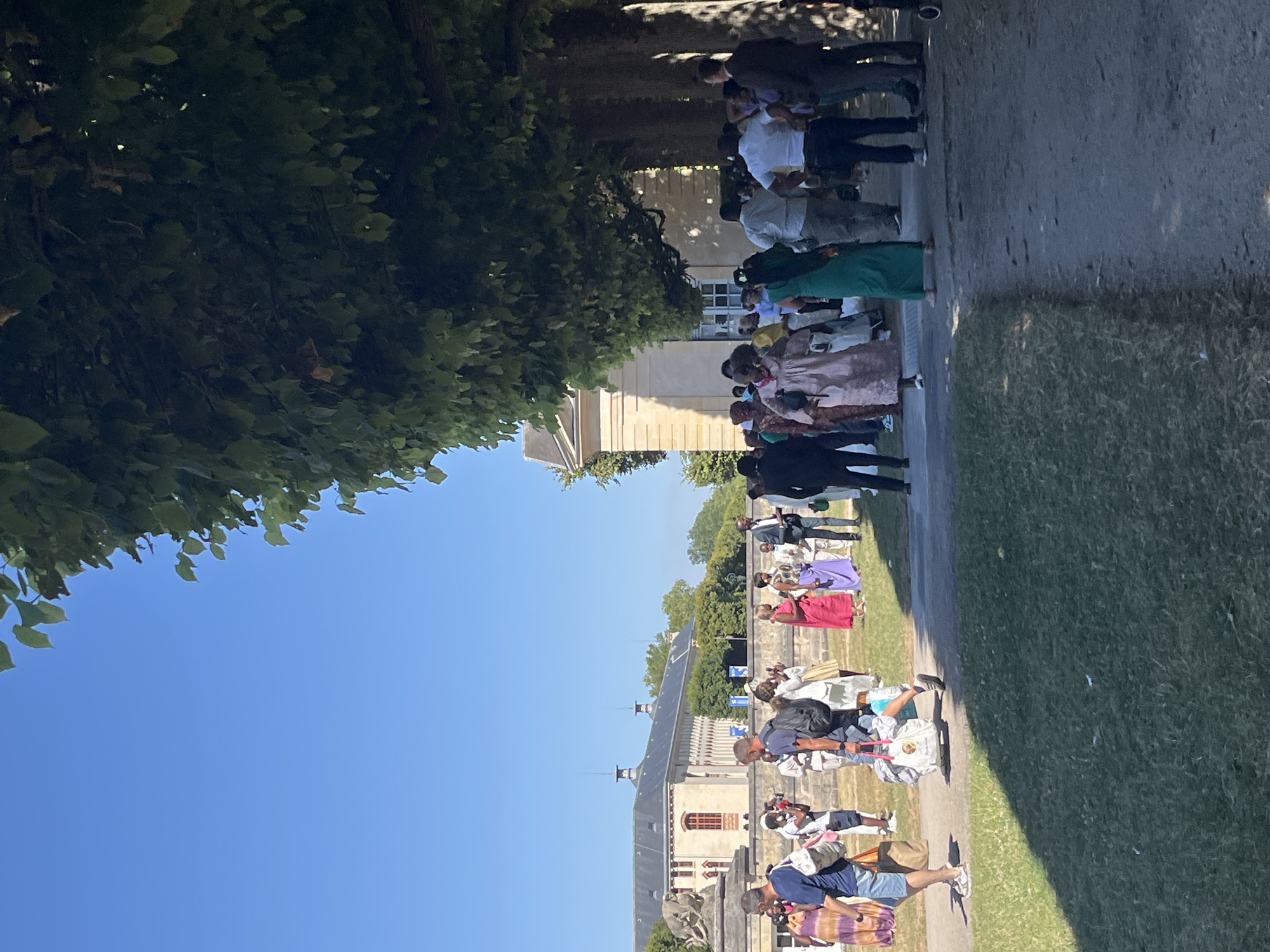 A group of people are gathered outdoors on a sunny day, on a path lined with trees in the city of Sceaux, France, with a historic building in the background. The building appears to be the Château de Sceaux, a large, neoclassical structure with a grey roof and light-colored walls, featuring arched windows and decorative elements. In the foreground, a gravel path runs diagonally from the lower right to the mid-left of the frame. To the left of the path, a grassy area is visible, partially shaded by the trees.
The people in the image are diverse in age and attire, with many wearing brightly colored clothing, some appearing to be of African origin. Several individuals are walking, while others are standing in small groups, suggesting social interaction or an event. One person in the foreground is carrying several bags. The trees lining the path cast dappled shadows on the ground. The sky is a clear, bright blue.