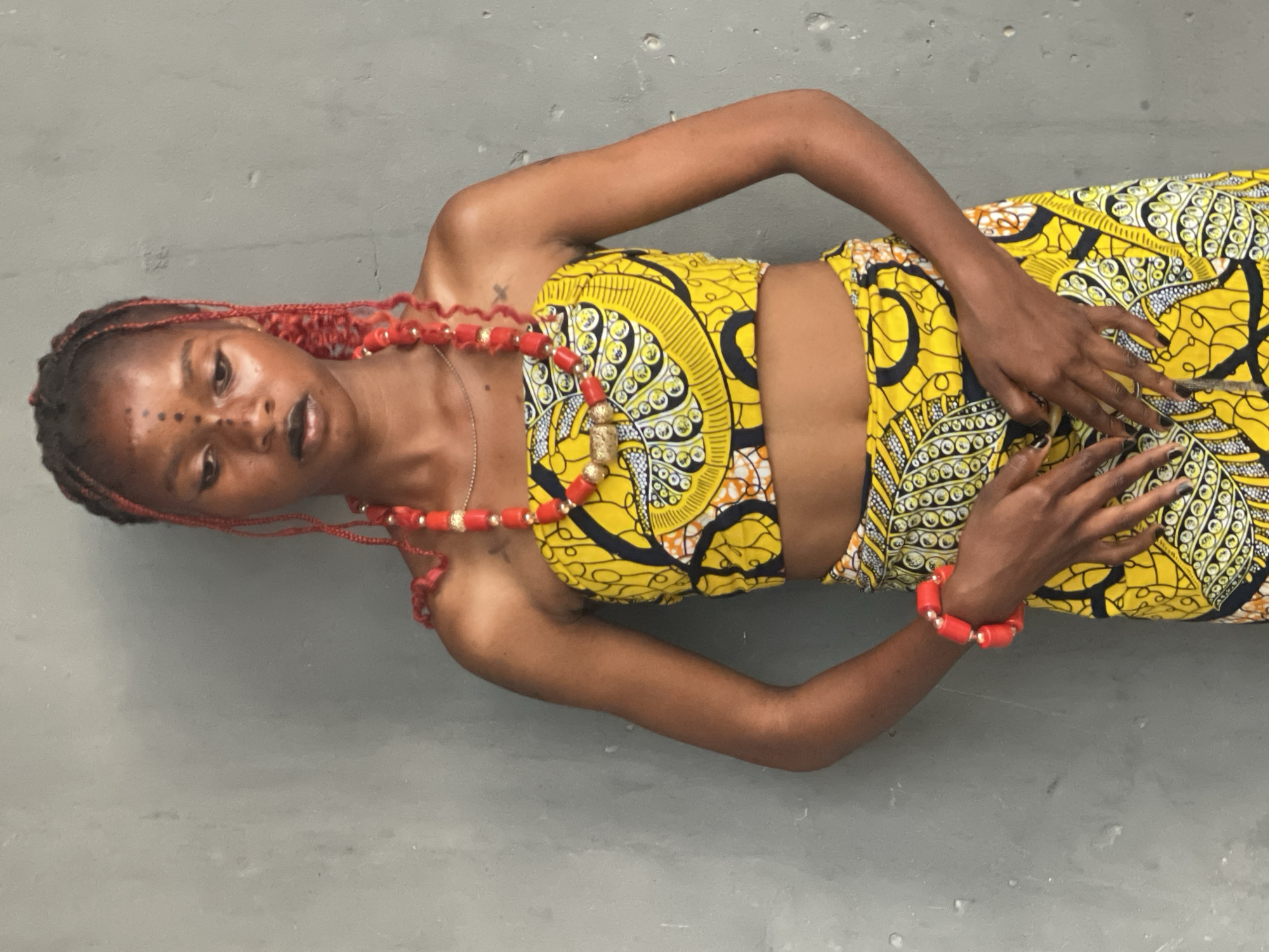 A young Black woman is depicted in a full-body portrait, lying on a plain grey surface, against a matching grey background. The setting is Badagry, Nigeria. She is dressed in a vibrant two-piece ensemble made from traditional African Ankara fabric, featuring bold yellow, black, and white circular patterns with hints of orange. The outfit comprises a strapless crop top and a matching skirt.
Her hair is styled in long, red braided extensions, some of which are embellished with small golden beads. Adorning her neck is a striking red beaded necklace with larger cylindrical red and golden beads, and she wears a coordinating red beaded bracelet on her right wrist. A delicate silver chain necklace is also visible. Her fingernails are painted black.
The woman has distinct black dot markings on her face: a line of small dots on her forehead and a cluster on the bridge of her nose. Her expression is calm and direct. Her hands are gently placed over her abdomen. A small, dark cross-like tattoo is visible on her left upper chest area. The lighting is even and soft, suggesting a daytime indoor setting. There is no visible text in the image.