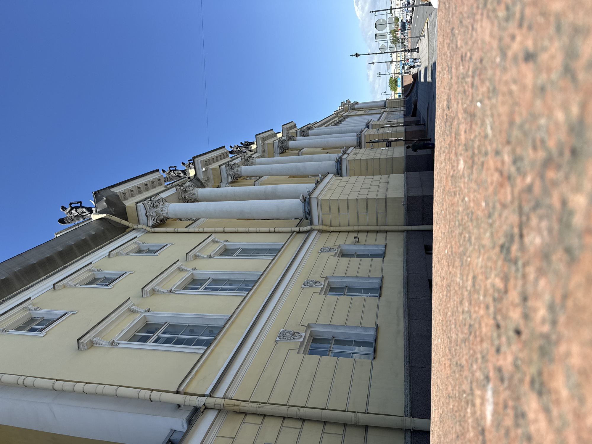 The image depicts a building facade in Saint Petersburg, Russia. The building features a pale yellow exterior with white trim, windows, and classical architectural elements such as columns. Gargoyles are visible along the roofline. A portion of a red granite embankment is in the foreground, partially obscuring the view. The sky is clear and blue. The image is taken from a low angle, looking upwards towards the building. People and other buildings are visible in the distance.
