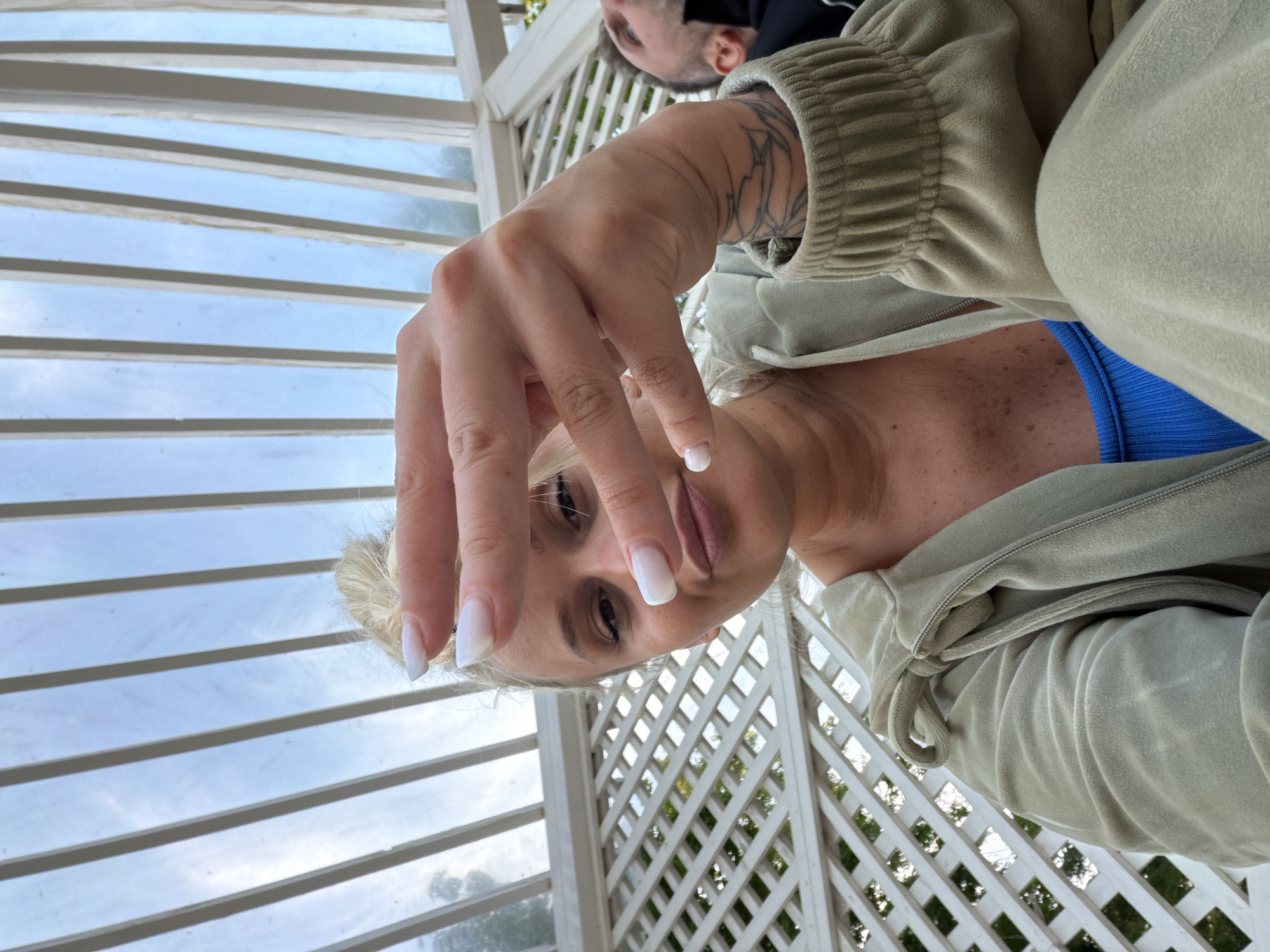 A woman with white-blonde hair and manicured nails is pictured from an unusual angle, likely taken by someone standing above her. Her left hand is prominent, with long, squared-off white nails. She is wearing a light green or beige jacket, and a glimpse of a blue garment is visible underneath. Her face is in focus, showing her eyes and part of her lips. A tattoo is visible on her left arm. In the background, there is a white structure, possibly a gazebo or a covered porch, with a lattice design and horizontal slats. A person is partially visible in the upper portion of the image, wearing a black face mask. Outside the white structure, greenery is visible, suggesting trees or foliage. The scene appears to be set outdoors, during the daytime. There is no indication that this image was taken in Sarapul, Russia.