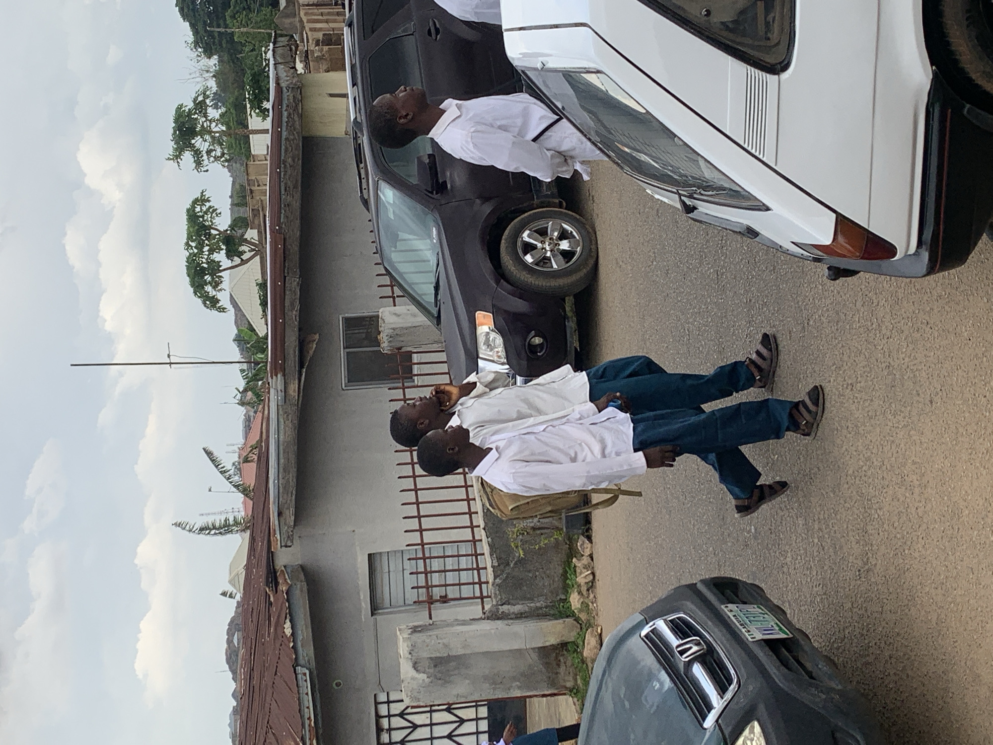 In Jos, Nigeria, two young men in white shirts and dark trousers walk on a street beside parked vehicles. The man on the right, with his hand raised to his face, appears to be speaking on a mobile phone. The man on the left carries a light brown backpack over his left shoulder. A dark-colored SUV is parked behind them, and a white hatchback is to their right. The background shows residential buildings with corrugated iron roofs and a cloudy sky. The street is paved, and there is a concrete wall with a metal fence in front of a building on the left. The image captures a moment in a typical urban environment in Jos.