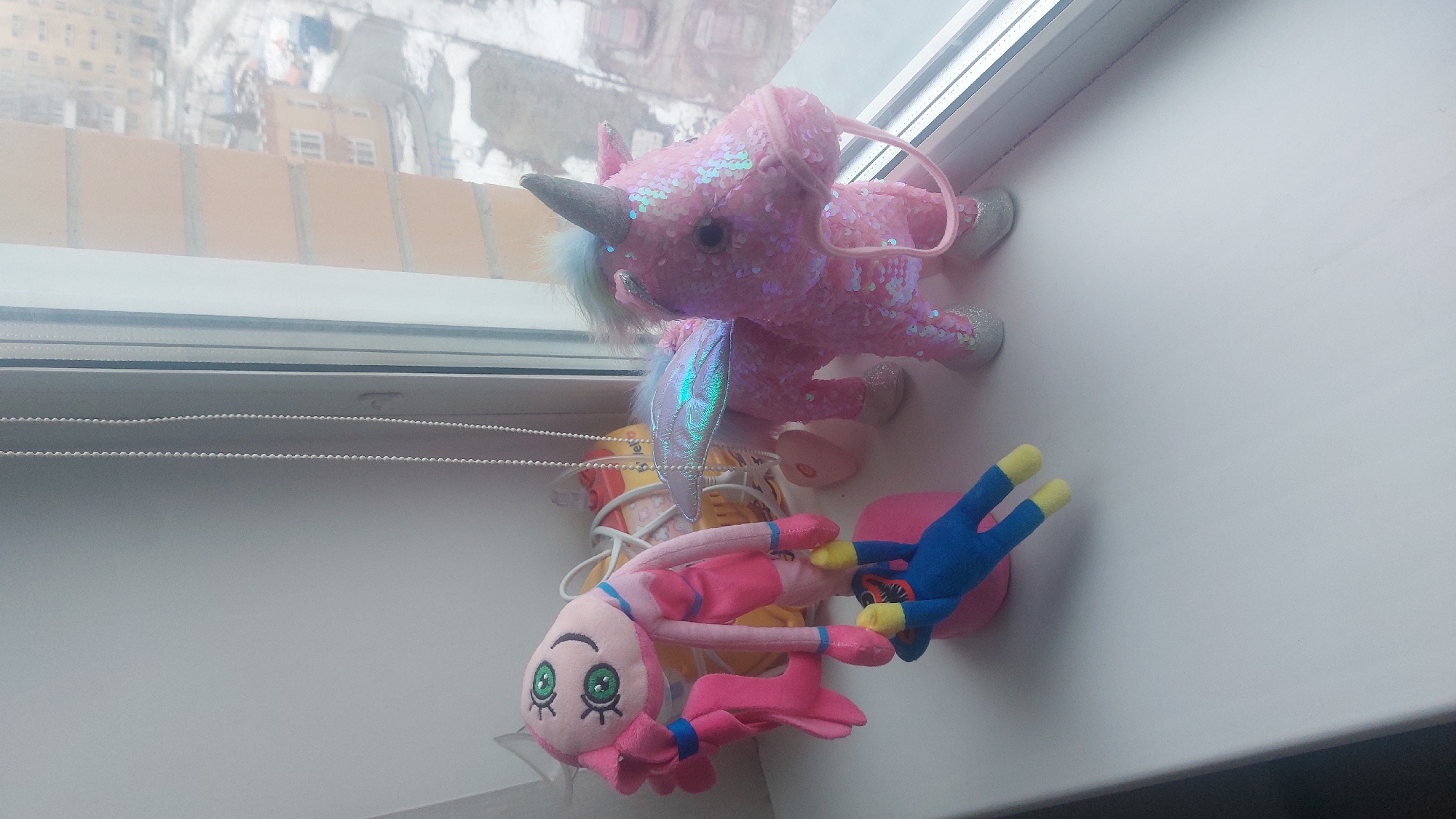 The image depicts a window sill in a room, presumably in a building in Novosibirsk, Russia, as suggested by the exterior view. The window is a modern double-glazed unit. On the window sill are two plush toys. The first toy is a pink, sequined unicorn with a silver horn and iridescent wings. The second toy is a pink, long-limbed character, possibly "Mommy Long Legs" from the "Poppy Playtime" game, coupled with a blue and yellow "Huggy Wuggy" plush. Between the toys is a partially visible yellow object, likely a toy or packaging. The room appears to be lit by natural light, with the exterior view showing a cityscape with buildings covered in snow. The window blinds are partially drawn.