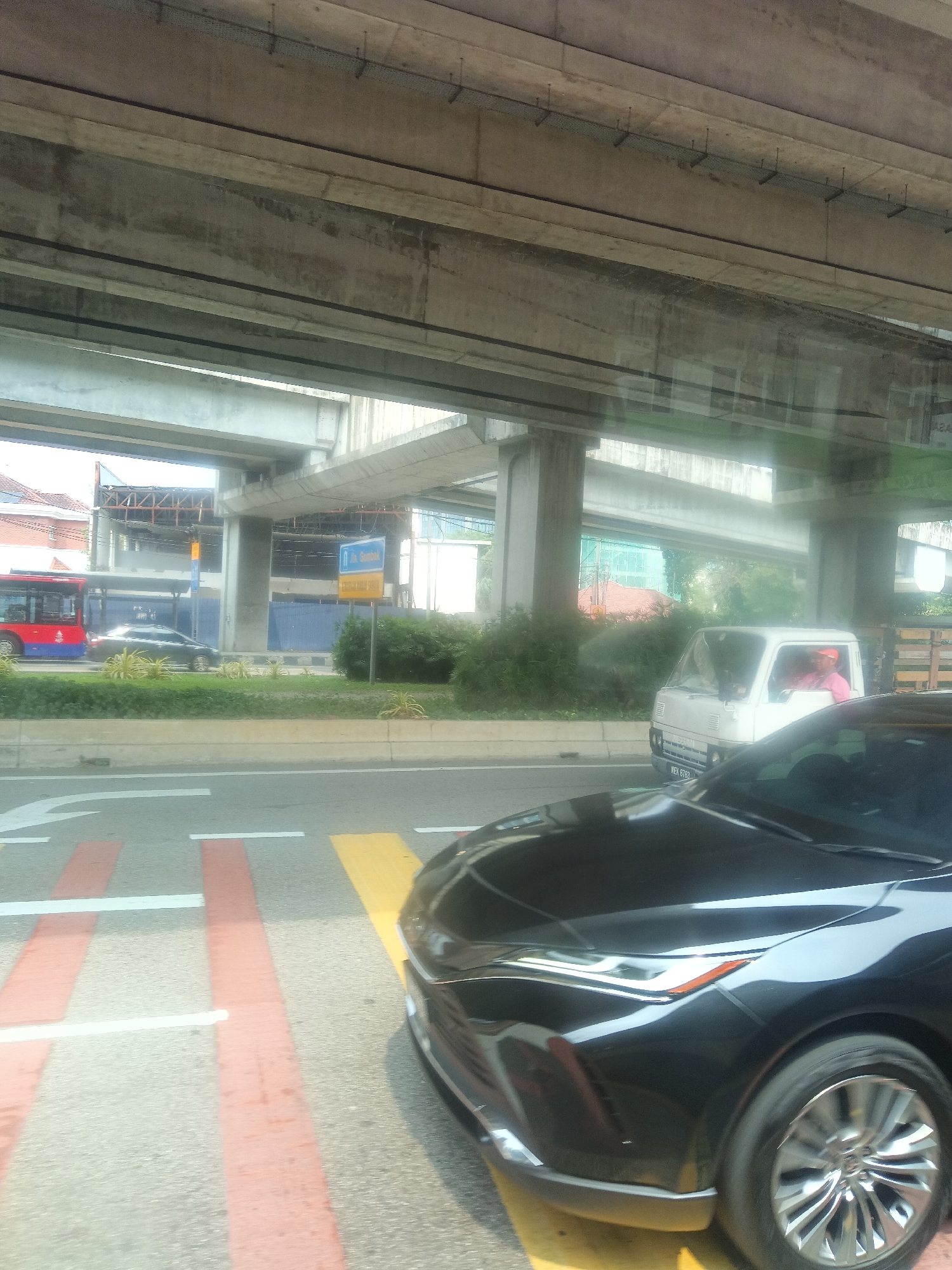 The image captures a street scene in Kuala Lumpur, Malaysia, under an elevated structure, likely a highway or an LRT line. In the foreground, the hood and headlights of a black car are visible, positioned on a road with pedestrian crossing markings. Further down the road, a dark-colored sedan is moving. To the left, a red and blue bus is parked or traveling. On the right side of the road, a small white truck with a driver wearing a pink shirt and an orange cap is also present. The background shows buildings under construction and greenery. A blue sign with text in Malay is visible, indicating "Jalan Gombak." The overall scene depicts urban traffic flow beneath a significant piece of city infrastructure.