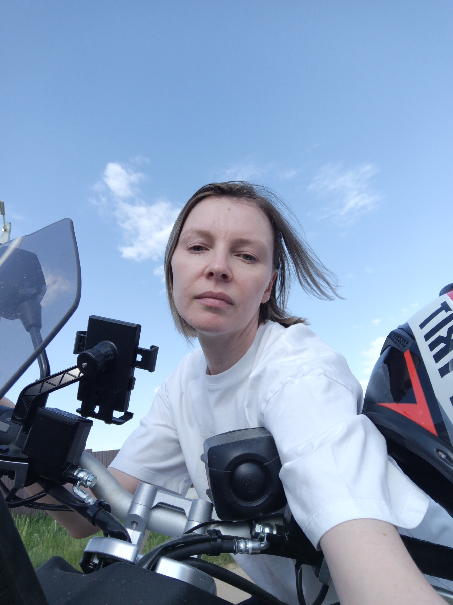 A medium, upward-angle shot features a light-skinned woman positioned on a motorcycle, likely outdoors, in what appears to be Chaikovsky, Russia.
The woman, facing the camera with a serious expression, has light brown hair that is wind-blown. She wears a white, short-sleeved shirt.
The motorcycle's handlebars are in the foreground, along with a black phone mount, brake fluid reservoir, and various cables. Part of a motorcycle helmet can be seen on the right, with red and white details.
In the background, a blue sky with scattered white clouds is visible. A fence is visible to the left.