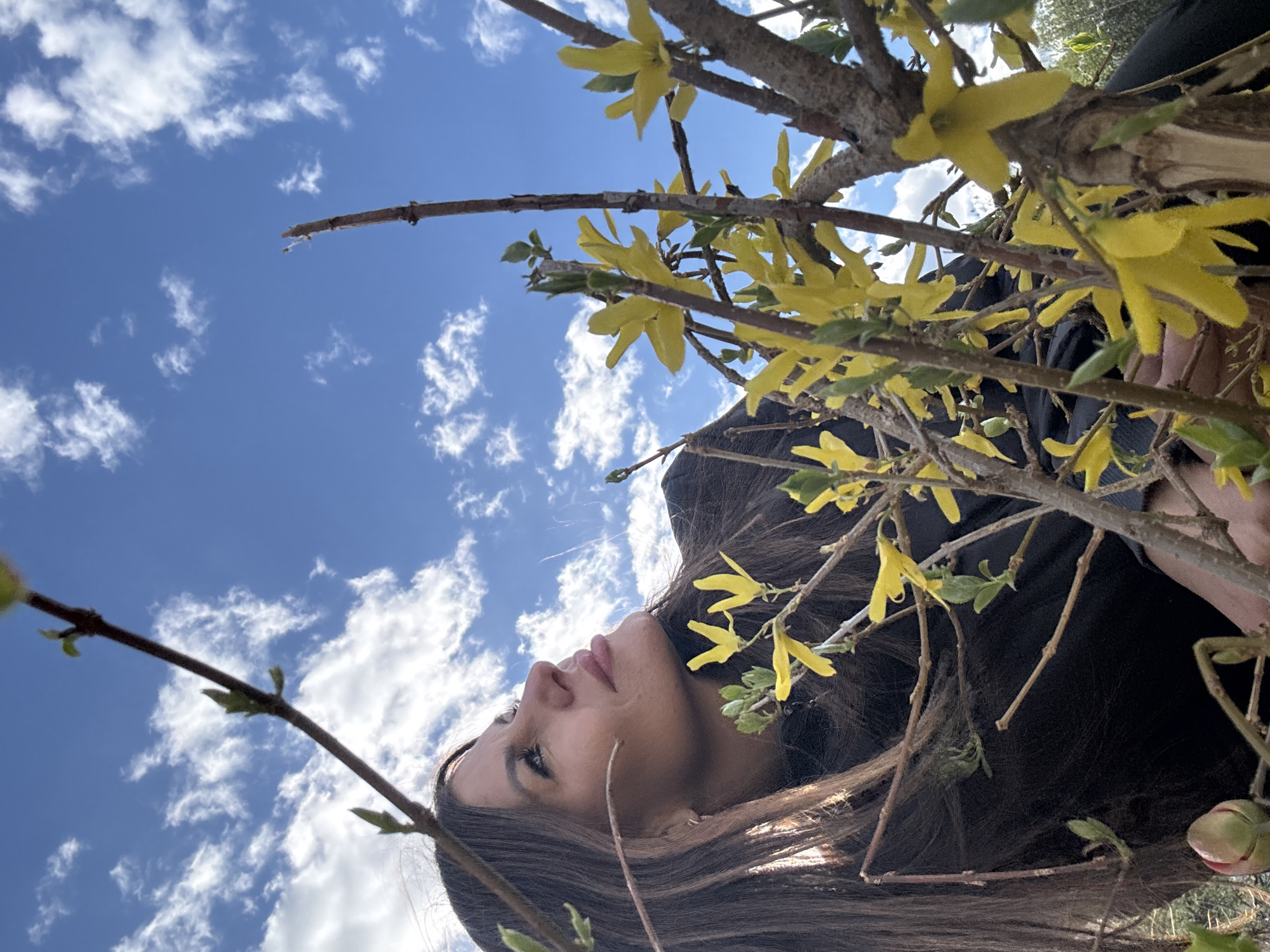 A woman is seen from a low angle, with the sky and clouds in the background. Branches with yellow flowers frame her face. The woman has long brown hair and is wearing a black shirt. The scene may be located in a natural setting, possibly a park or garden. The city of Dubovaya, Russia is not discernible from the image.