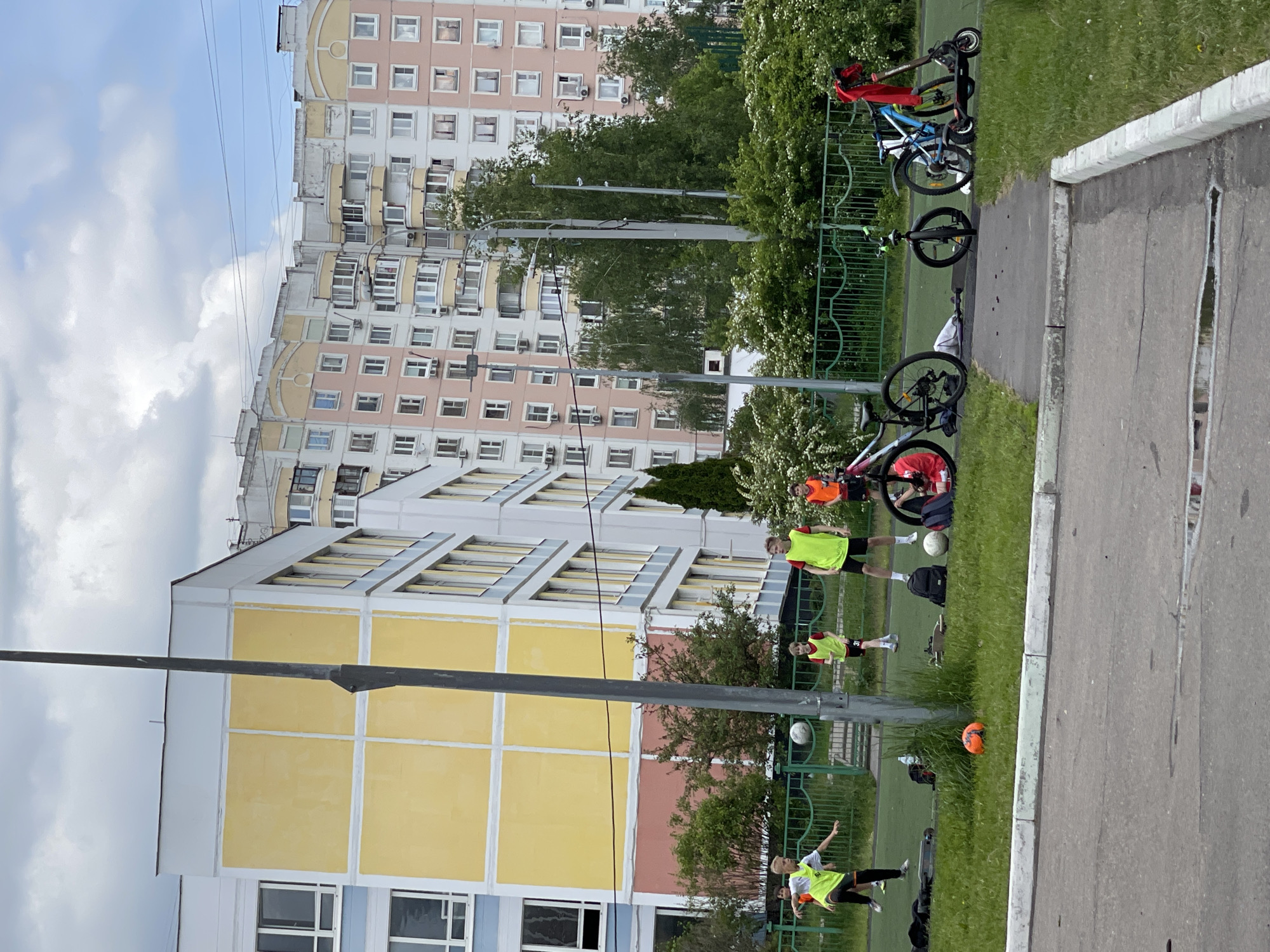 The image shows a group of children playing football in a courtyard in Moscow, Russia. The location is identifiable by the typical architecture of Soviet-era apartment buildings in the background. The children are wearing yellow vests. Some are kicking or about to kick a football, while others are standing and observing the game. Bicycles and scooters are parked near the green fence. Backpacks and other objects are on the grass nearby. The ground is partly grass and partly paved. Overhead wires cross the sky in the background.