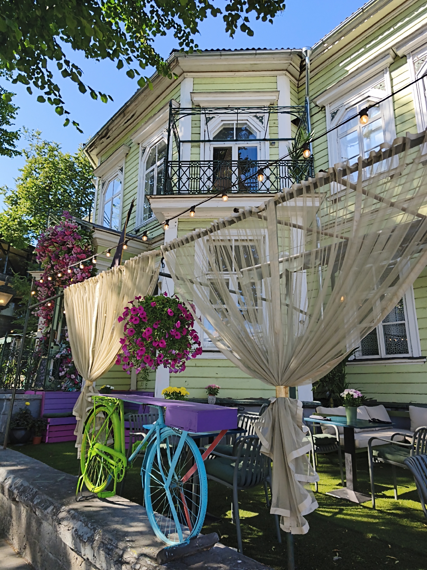 This image captures a vibrant outdoor seating area in front of a light green wooden building, likely a cafe or restaurant, situated in the historic Old Town of Tallinn, Estonia. The building features ornate white trim around windows and a second-story balcony with a wrought-iron railing. String lights with exposed bulbs are draped across the scene, adding a festive atmosphere. Sheer beige curtains hang from a metal frame, partially obscuring the view of the building's facade and creating a whimsical, layered effect.
In the foreground, two vibrantly painted bicycles, one lime green and the other bright blue, are positioned as decorative elements against a rough stone wall. The green bicycle is leaning against a purple structure, and the blue bicycle has its front wheel turned outward. Behind the bicycles, a low purple bench is visible, adorned with potted plants.
The seating area itself consists of outdoor tables and chairs set up on a well-maintained lawn. A glass-topped table with dark metal legs is surrounded by chairs, and further back, cushioned seating arrangements are visible, suggesting a comfortable place for patrons to relax. Lush pink and purple flowers cascade from hanging baskets and arrangements, adding splashes of color and enhancing the romantic ambiance. The presence of flowers, decorative bicycles, and outdoor seating implies this is a popular spot for visitors to enjoy refreshments and the charming atmosphere of Tallinn's Old Town. The image is taken from a slightly low angle, looking up towards the building, with the dappled sunlight filtering through green leaves from a tree overhead.
