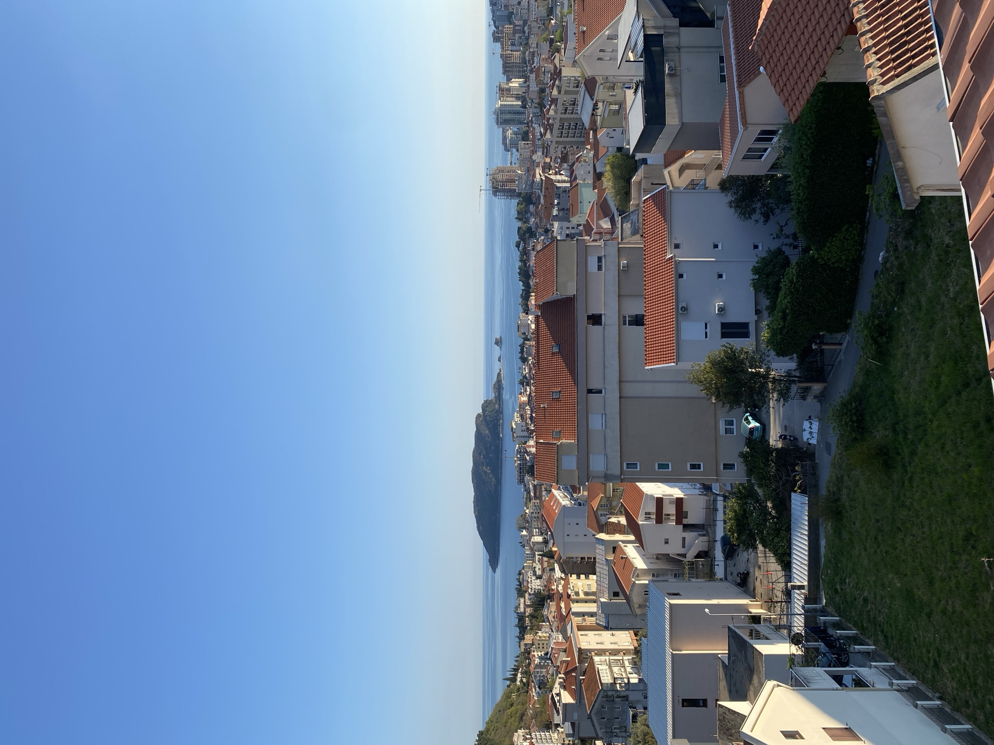 A vertically oriented, high-angle outdoor shot depicts the coastal city of Budva, Montenegro, under a clear blue sky. The Adriatic Sea stretches along the horizon, with a long, verdant island situated parallel to the coastline. The shoreline is densely populated with buildings, mainly residential, with terra-cotta roofs and light-colored facades. Some greenery, including trees and grassy areas, is interspersed among the buildings. A street can be seen winding through the residential area. A few parked cars are visible. A building rooftop occupies the lower right corner of the image.