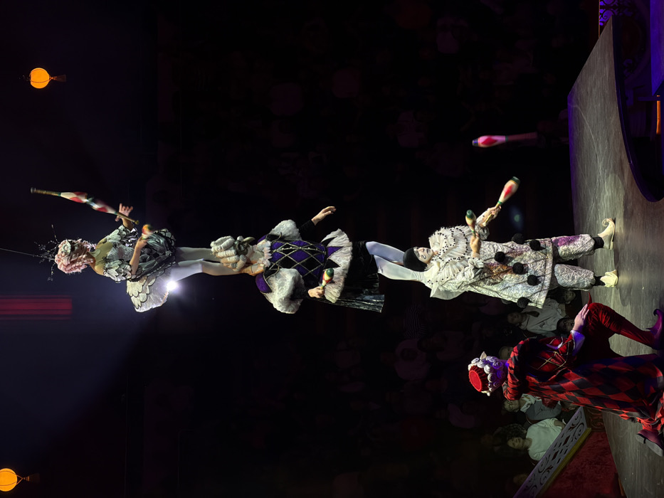 This image captures a multi-person acrobatic and juggling performance inside a large circus venue in Moscow, Russia.
The central focus is a three-person human pyramid. The base performer, dressed in a white patterned jumpsuit with black pom-poms, a ruffled collar, white face paint, and a black cap, stands on a dark stage. This individual supports a second performer on their shoulders. The second performer wears a purple and black harlequin-patterned tunic with white ruffles and a large white powdered wig. This performer, in turn, supports a third, top performer on their shoulders. The top performer, a female acrobat, is clad in a white ballet-style tutu with silver-grey embellishments, white tights, and a complex headpiece featuring pink hair and a mask-like facial covering. She balances on one leg. Both the top and the base performers are actively juggling pins; one pin is blurred mid-air near the base performer, and another is blurred in motion near the top performer.
To the lower left, a fourth performer kneels on one knee on a slightly raised, ornate red platform. This individual wears a red and black diamond-patterned cloak, matching tights, and a red cap with a white textured top, positioned with their back partially to the central acrobats.
The stage is dimly lit by spotlights directed at the performers. The background shows blurred, darkened seating, indicating an audience in a large arena. Two spherical, orange-yellow lanterns are suspended in the upper corners of the frame.