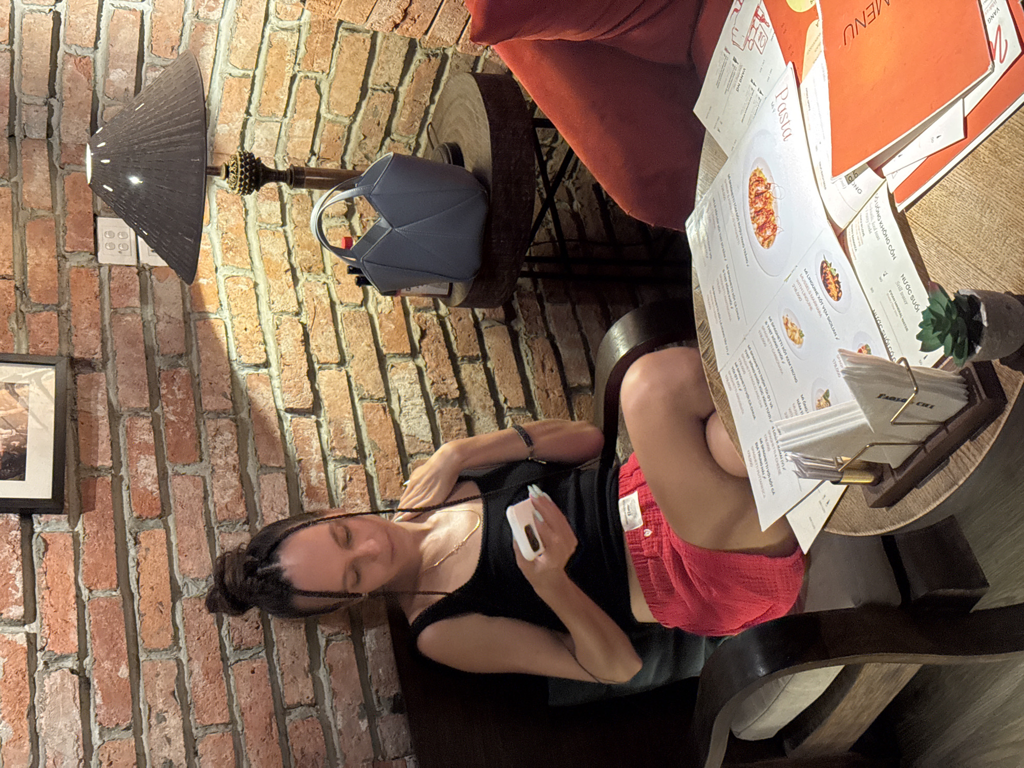 A woman sits in a wooden armchair in a restaurant with exposed brick walls. She is wearing a black tank top and red shorts. She is holding a white phone in her hands and looking down at it. In front of her is a table with menus and a small plant. A gray handbag is visible on a side table. The lighting is dim, suggesting an indoor setting possibly at night or in a dimly lit cafe. The menus appear to have text in Vietnamese. The brickwork is prominent, and there's a framed picture on the wall above the woman. A red sofa is partially visible to the right of the frame.