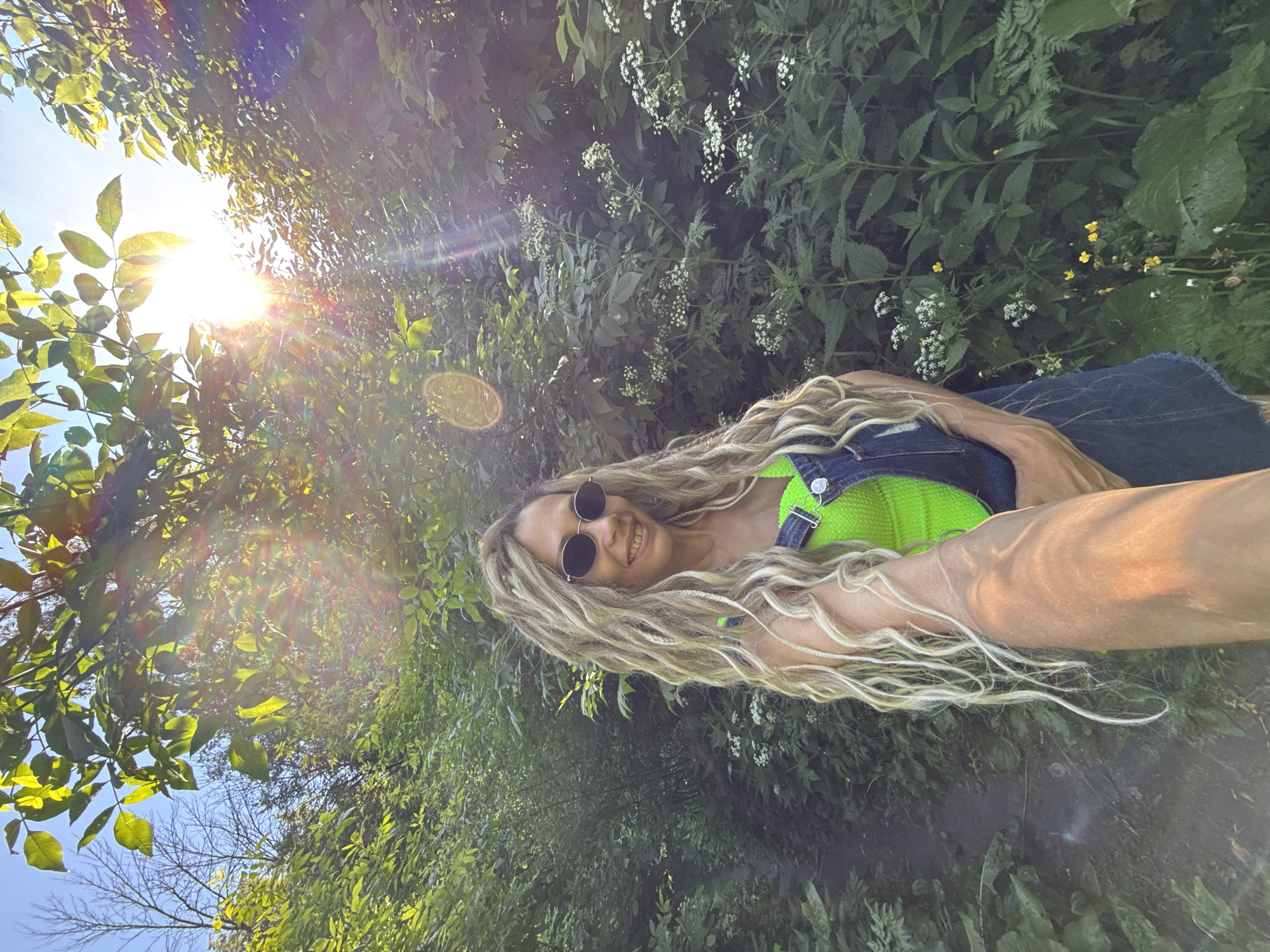 In a sun-dappled forest setting, likely in the vicinity of Шебунино, Russia, a woman with blonde dreadlocks and sunglasses poses for a selfie. She is wearing a bright neon green top and denim overalls. The lush green foliage of trees and plants surrounds her, with sunlight streaming through the leaves, creating a lens flare effect. A dirt path is visible to her left, suggesting a trail or walking path within the forest. The overall scene conveys a sense of nature and outdoor recreation.