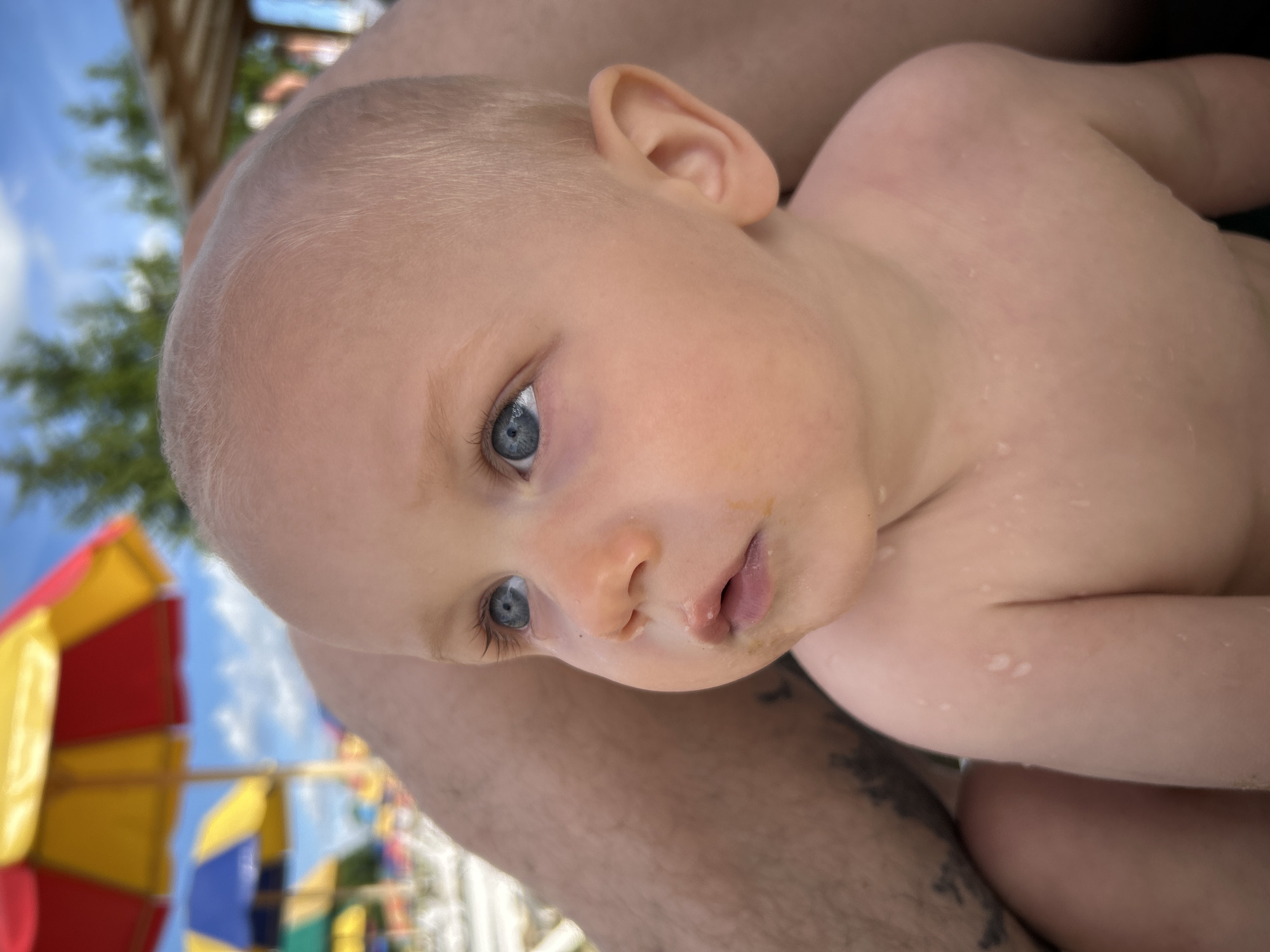 A close-up, tilted shot focuses on the face of a young child with light hair and striking blue eyes. The child is held by an adult, whose arm is visible in the foreground. The child's skin has a few droplets of water, suggesting they may have been swimming or playing in water. They have a neutral expression, with their gaze directed slightly away from the camera.
In the background, out of focus, are colorful elements that resemble parts of a playground or amusement area. There are large, umbrella-like structures with bright red, yellow, and blue sections. The sky is visible, appearing to be a clear blue with some white clouds. The overall setting seems to be outdoors, possibly at a water park or a recreational area on a sunny day.
The image is oriented such that the child's head is at the top right, and the adult's arm and the background elements extend towards the bottom left. There is no visible text in the image. The time of day appears to be daytime, based on the bright lighting and clear sky.