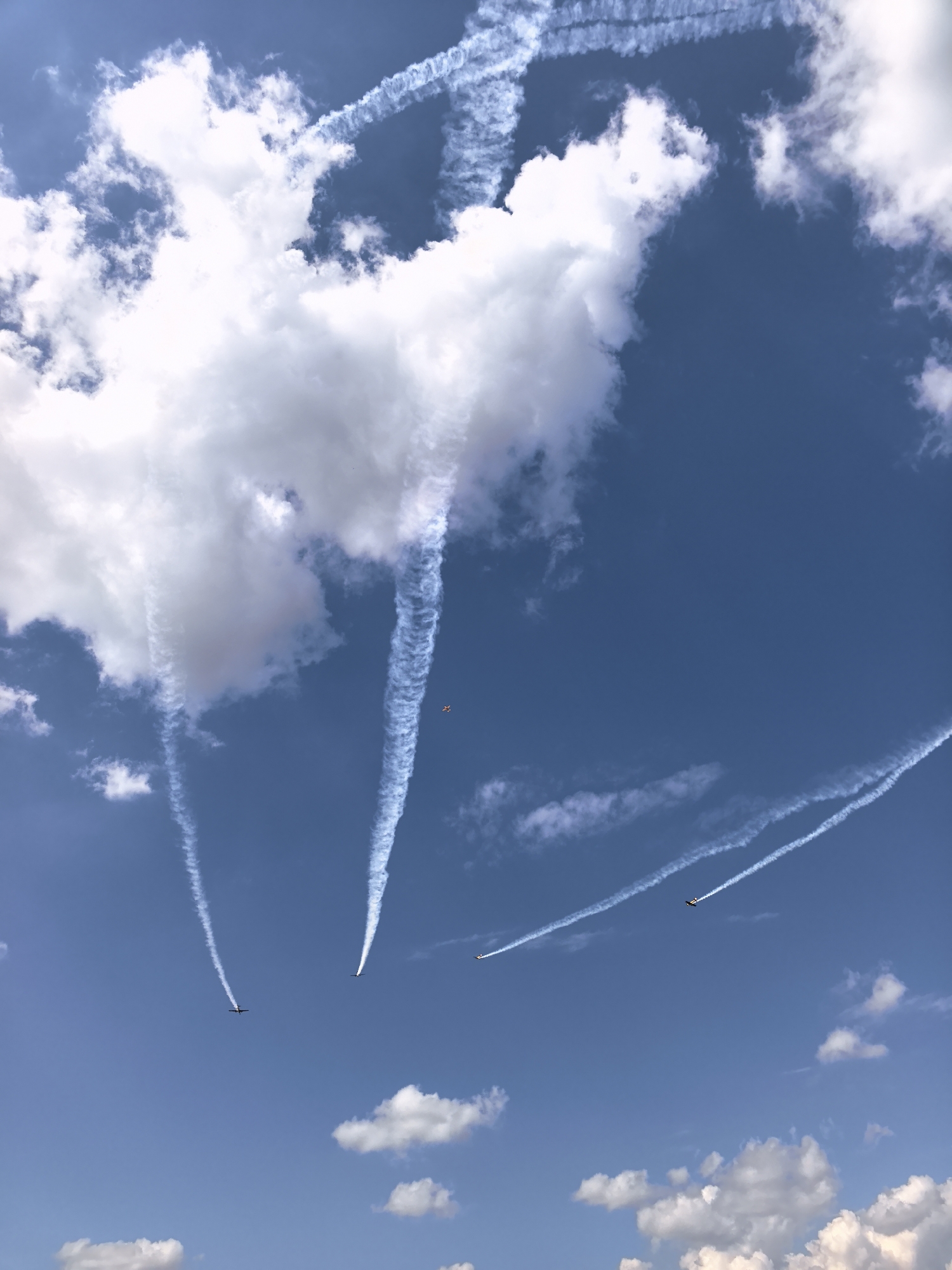 The image captures an aerial performance against a bright blue sky with scattered white clouds. Several planes are visible, leaving trails of white smoke. Some of these trails form intricate patterns, suggesting aerobatic maneuvers. One small, distant plane is colored orange and white. The overall scene is clear and detailed, suggesting it was taken on a sunny day. The specific location in Nestiukovo, Russia, is not visually discernible from the image itself.