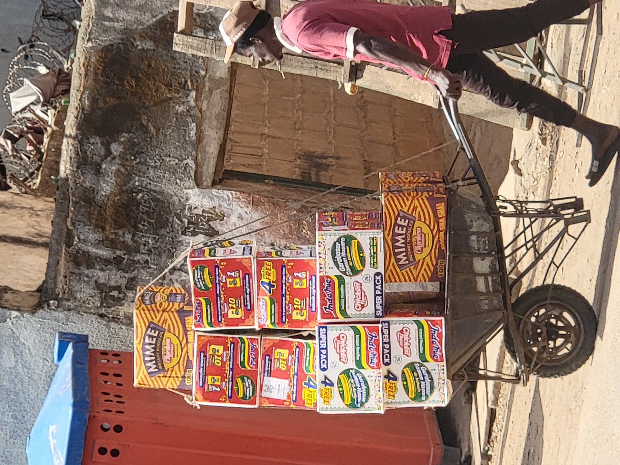 A man in a maroon shirt, dark trousers, and a beige cap is depicted pushing a metal handcart laden with instant noodles in a street or market setting in Maiburiji, Nigeria. His posture suggests he is actively engaged in moving the heavily loaded cart forward.
The cart is stacked high with numerous cardboard boxes of instant noodles. Two main brands are prominently visible: "MIMEE INSTANT NOODLES" in distinctive yellow and orange packaging, and "Indomie Chicken" in red, white, and green boxes. Many boxes display promotional text such as "SUPER PACK" and "4 FREE," while others include "Garabasar Somun Nasaro." Additional text like "10" and "100g" indicates quantity or weight.
The background consists of a textured, light-colored wall, possibly concrete or mud, showing signs of weathering and some dark stains. A wooden structure, resembling a shutter or door, is visible behind the man. The ground is dusty and light-colored. In the lower left foreground, part of a blue and red plastic container is visible. The bright lighting and distinct shadows indicate a sunny day, likely during the daytime. The scene captures a typical daily activity of goods transport in a local environment.