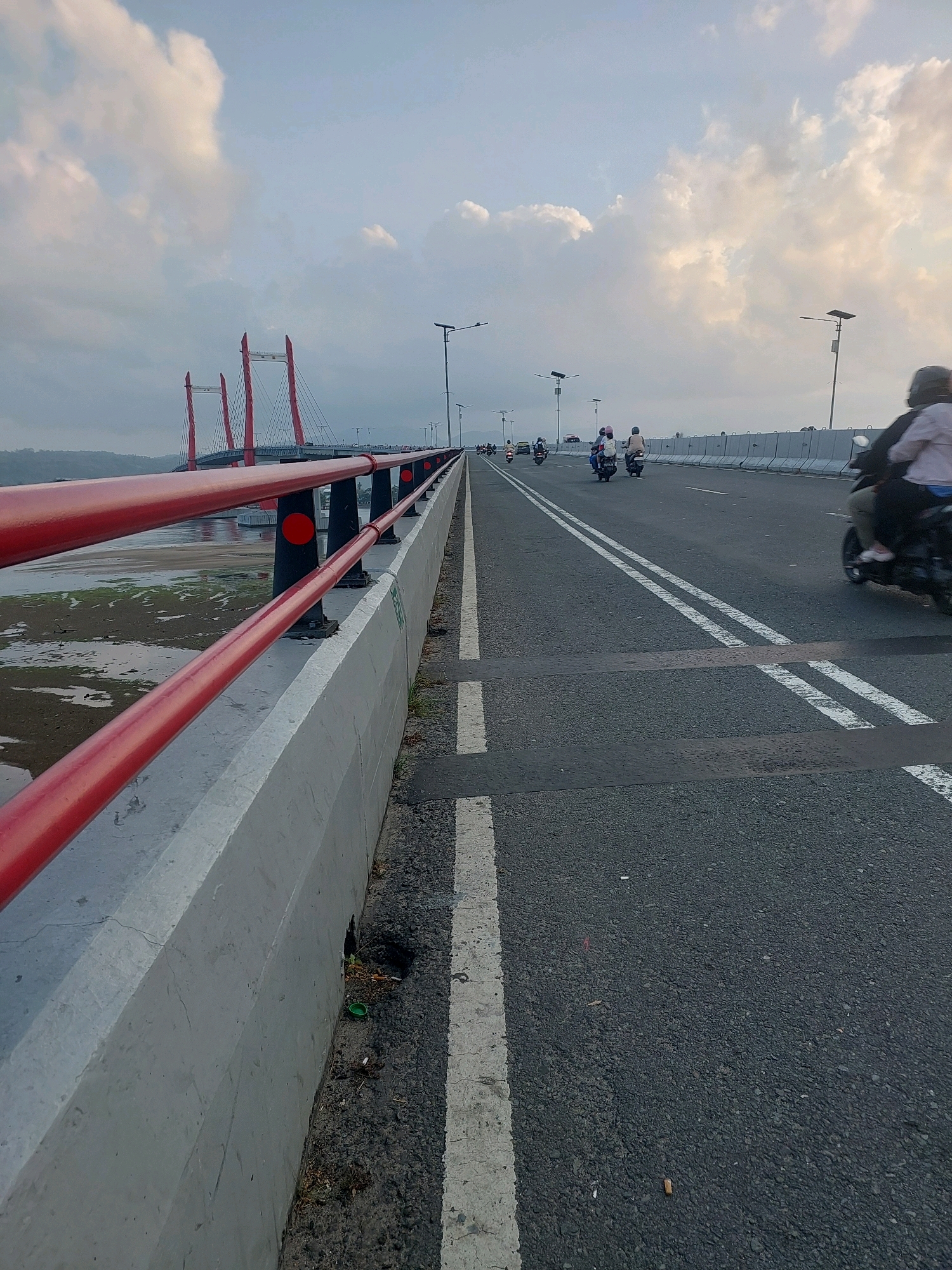 The image depicts the Poka-Galala Bridge in Ambon, Indonesia, during what appears to be late afternoon or early evening. The bridge is a cable-stayed bridge with prominent red pylons supporting the cables. The roadway is asphalt, marked with white lines, and traffic flows in both directions. Several motorcycles are visible on the bridge, some carrying passengers. The sky is partly cloudy, with soft, diffused light. On the left side of the bridge, a concrete barrier is topped with a red railing. Below the bridge, a muddy or sandy area with some water is visible, suggesting a tidal area or riverbank. Streetlights are spaced along the bridge. Some debris, including cigarette butts and a green bottle cap, is present on the ground near the concrete barrier. The overall scene is one of everyday activity on a significant piece of infrastructure.