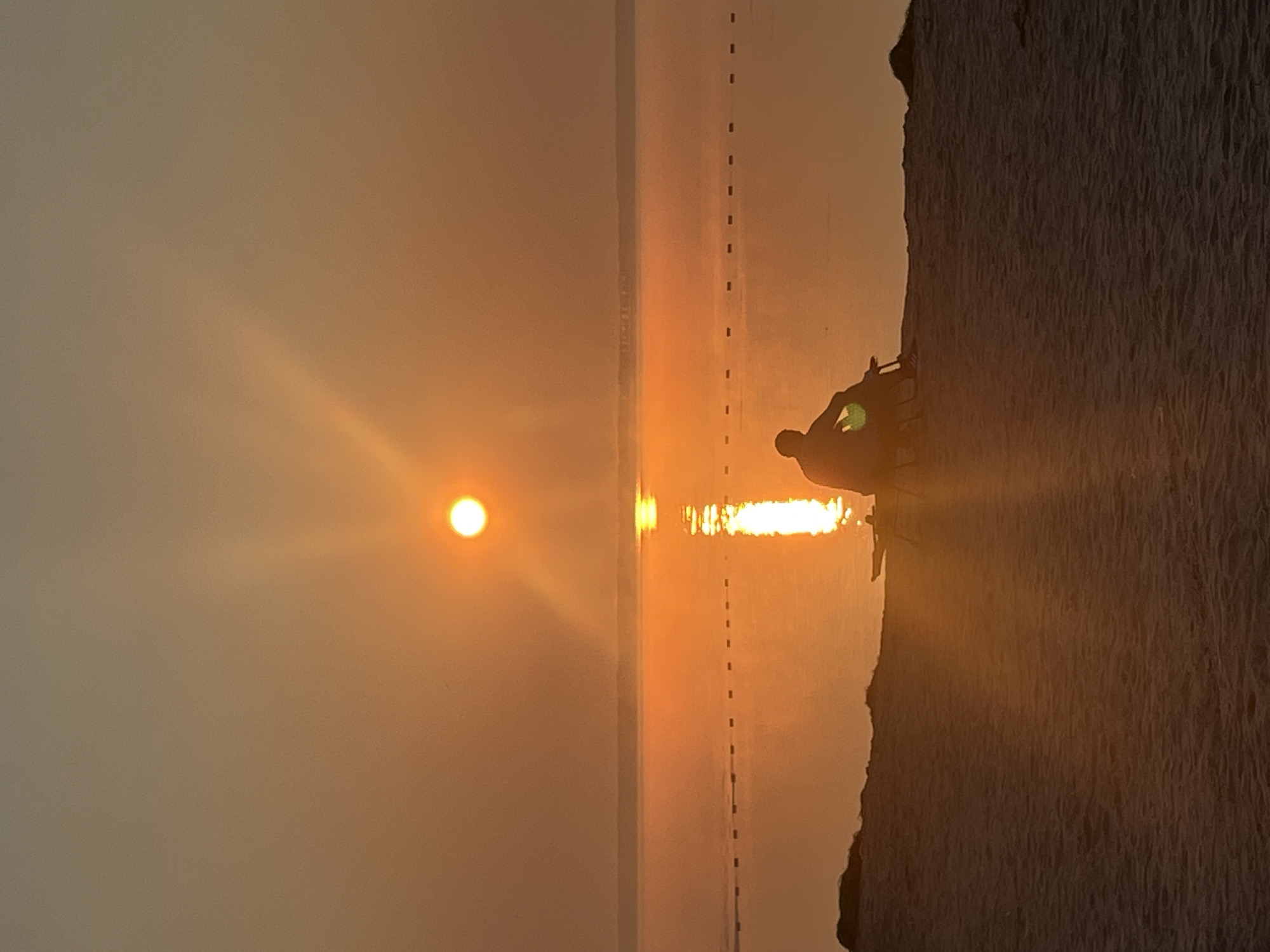 The image depicts a serene sunset scene on a sandy beach in Surakovo, Russia. The sun, a bright orb, hangs low in the sky, casting a warm, golden glow across the water and the horizon. Its reflection shimmers on the calm surface of the sea, creating a luminous path that stretches towards the shore. In the foreground, a person is silhouetted against the vibrant sunset, sitting on a beach chair on the sandy embankment. The person is facing the water, appearing to be in quiet contemplation. A few dark, indistinct objects, possibly rocks or vegetation, are scattered along the edge of the beach. The overall atmosphere is tranquil and peaceful, capturing a moment of solitude amidst the breathtaking natural beauty.