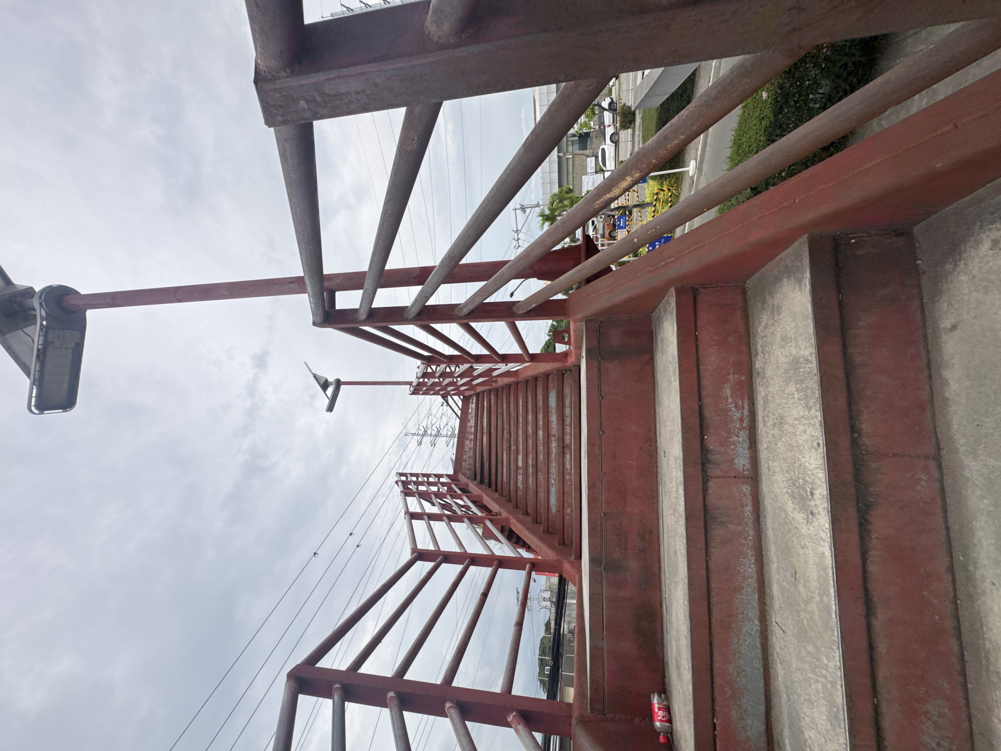 The image presents a view looking along a long pedestrian overpass constructed of concrete steps and rusty red metal railings and supports. The structure appears aged with visible weathering.
On the left side of the overpass, a street light fixture is attached to a pole. Below and to the left, a road is visible with multiple vehicles, including white vans, parked or moving. Adjacent to the road are low-rise buildings, some with awnings, and patches of green bushes.
Further down the overpass and to the right, a network of power lines and a large metal electricity pylon are visible against the sky, indicating an industrial or utility area. The sky overhead is overcast with light grey clouds, suggesting a daytime setting.
A discarded red and white plastic bottle, clearly labeled "Coca-Cola," lies on one of the concrete steps of the overpass. The scene depicts an urban or suburban environment in Davao City, Philippines. No people are visible in the image.