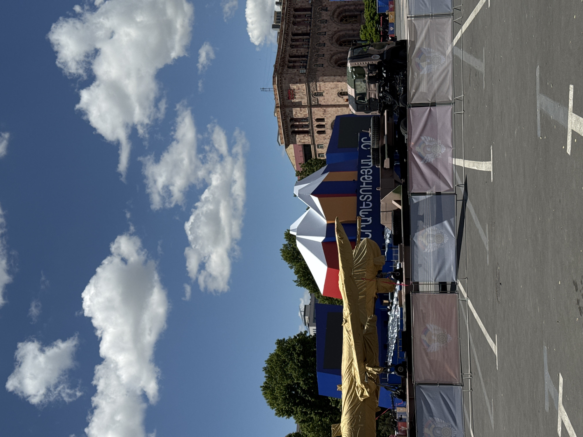 The image shows a section of Republic Square in Yerevan, Armenia. The view is looking toward the Government House building. A military parade is being set up, with covered statues, banners, and military vehicles present in the square. The sky is blue with scattered clouds.