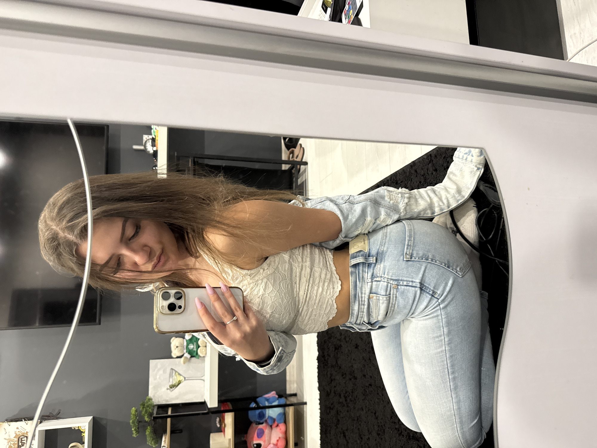 A woman is taking a selfie in a mirror. She is wearing light blue jeans, a white lace top, and a light blue denim jacket. The background is a room with a black rug and a shelf with various items. The location is not possible to determine from the image.