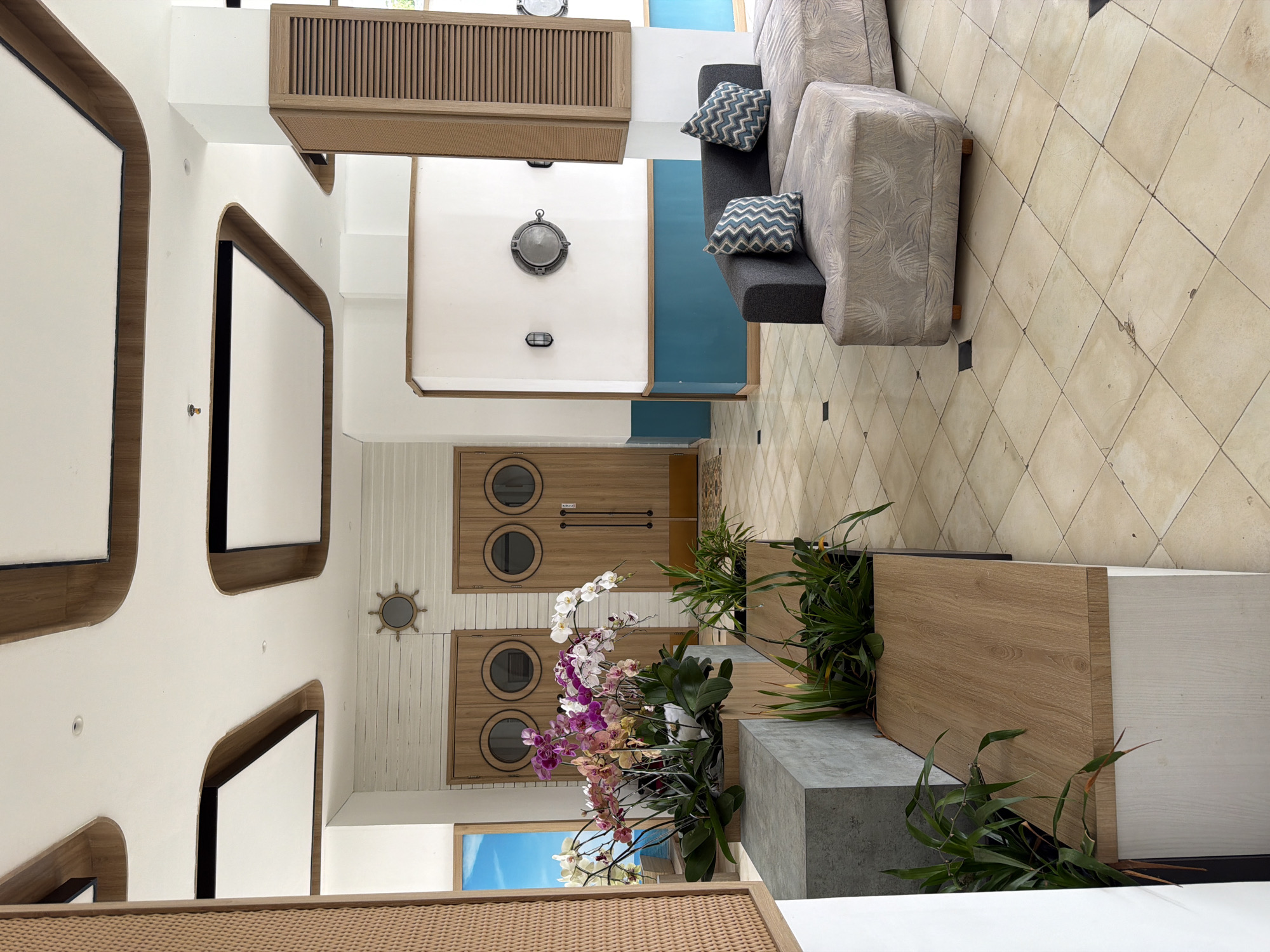 The image depicts the interior of a modern establishment, possibly a hotel lobby or a lounge area, with a nautical theme. The scene is well-lit and presents a clean, contemporary aesthetic.
Dominating the foreground on the left is a raised planter containing an abundance of colorful orchids and other greenery, arranged in geometric planters. To the right of the orchids, a plush sectional sofa with patterned upholstery and decorative blue and white zigzag pillows sits on a tiled floor. The floor itself is composed of large, square beige tiles with occasional darker square accents.
In the background, two large wooden doors with circular porthole-style windows are visible, suggesting a maritime or coastal influence. Above these doors, a ship's wheel emblem is mounted on a white, wood-paneled wall. Further to the right, a white wall features a large, circular porthole window and two wall-mounted light fixtures. A wooden louvered cabinet is positioned in the corner, adding a textural element. The ceiling is white with intricate wooden trim and recessed square lighting elements.
The overall impression is one of a stylish and inviting space that blends modern design with a nautical aesthetic. The location is identified as Unknown, Indonesia. There are no people visible in the image, and no discernible actions or interactions are taking place. The environment appears to be an indoor public space.