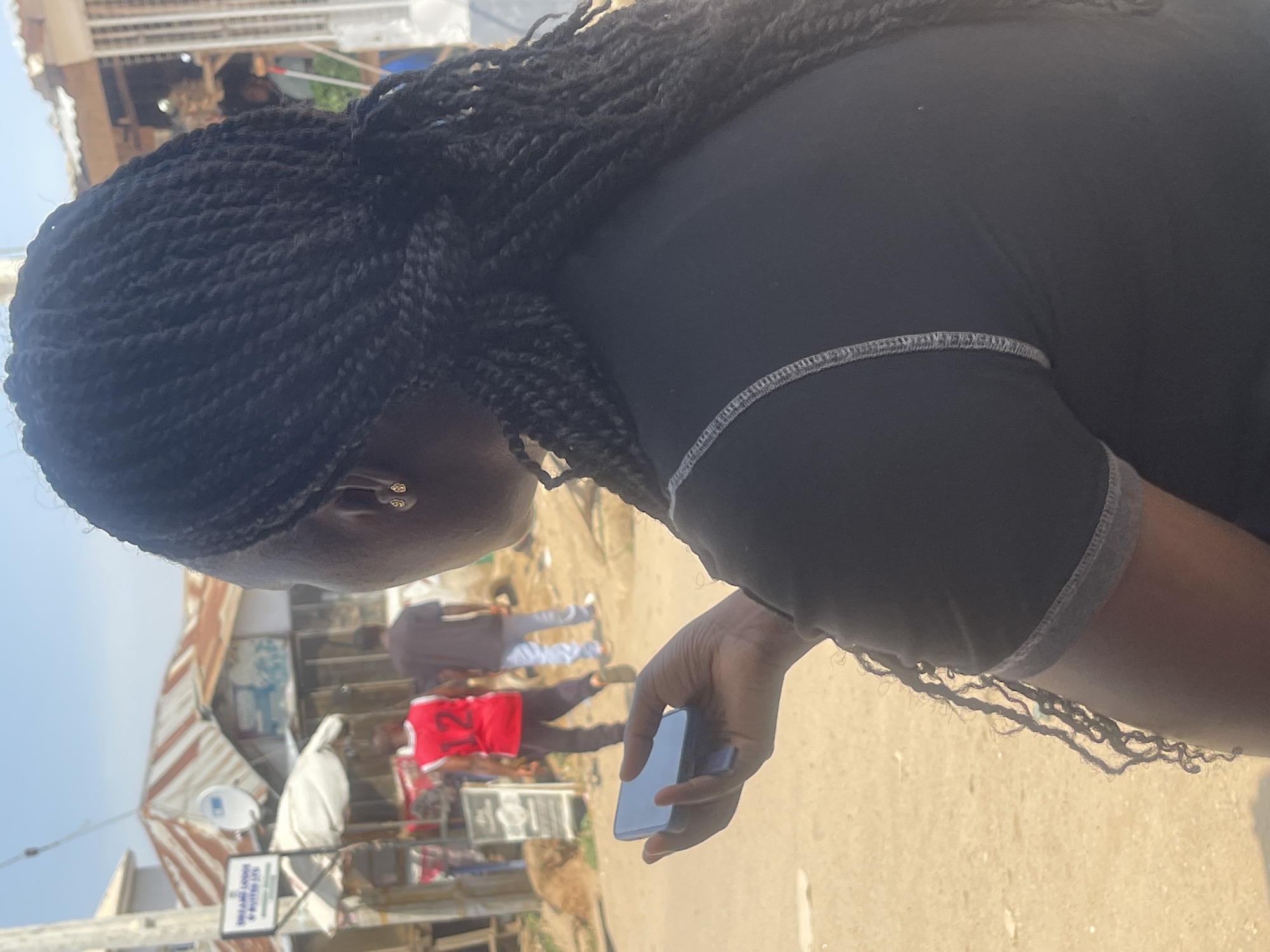 In Abuja, Nigeria, on a bright, sunny day, a woman with dark, braided hair is seen from behind, holding a dark-colored smartphone in her left hand. She is wearing a black t-shirt with grey stitching along the neckline and sleeves. Her right ear, adorned with a small gold earring, is visible. In the background, two men are walking away from the camera. One man is wearing a red sleeveless shirt with the number "12" on it and black pants. The other man is wearing a dark t-shirt and light-colored pants. They are on a dirt road in front of a row of buildings with corrugated metal roofs, suggesting a bustling market or residential area. A sign with text, partially obscured by a white canopy, is visible on the left side of the image. The environment appears to be an outdoor, urban setting.