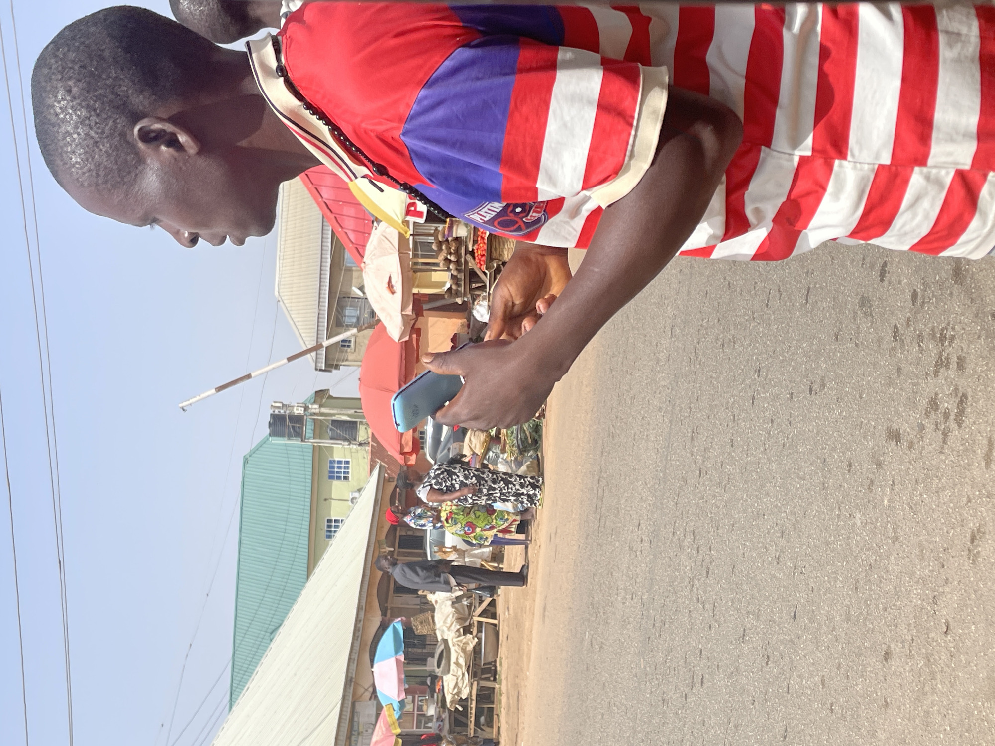 The image shows a man in a red and white striped shirt with blue shoulder panels standing in what appears to be an outdoor market area. The man is holding a smartphone and looking down at it. In the background, people are gathered at market stalls covered by various umbrellas and awnings. There's a paved road in the foreground, and buildings with corrugated roofs are visible in the mid-ground. The image is taken during daylight hours, and based on the context clues, it could be plausibly located in an urban area in Jos, Nigeria, where outdoor markets are common.