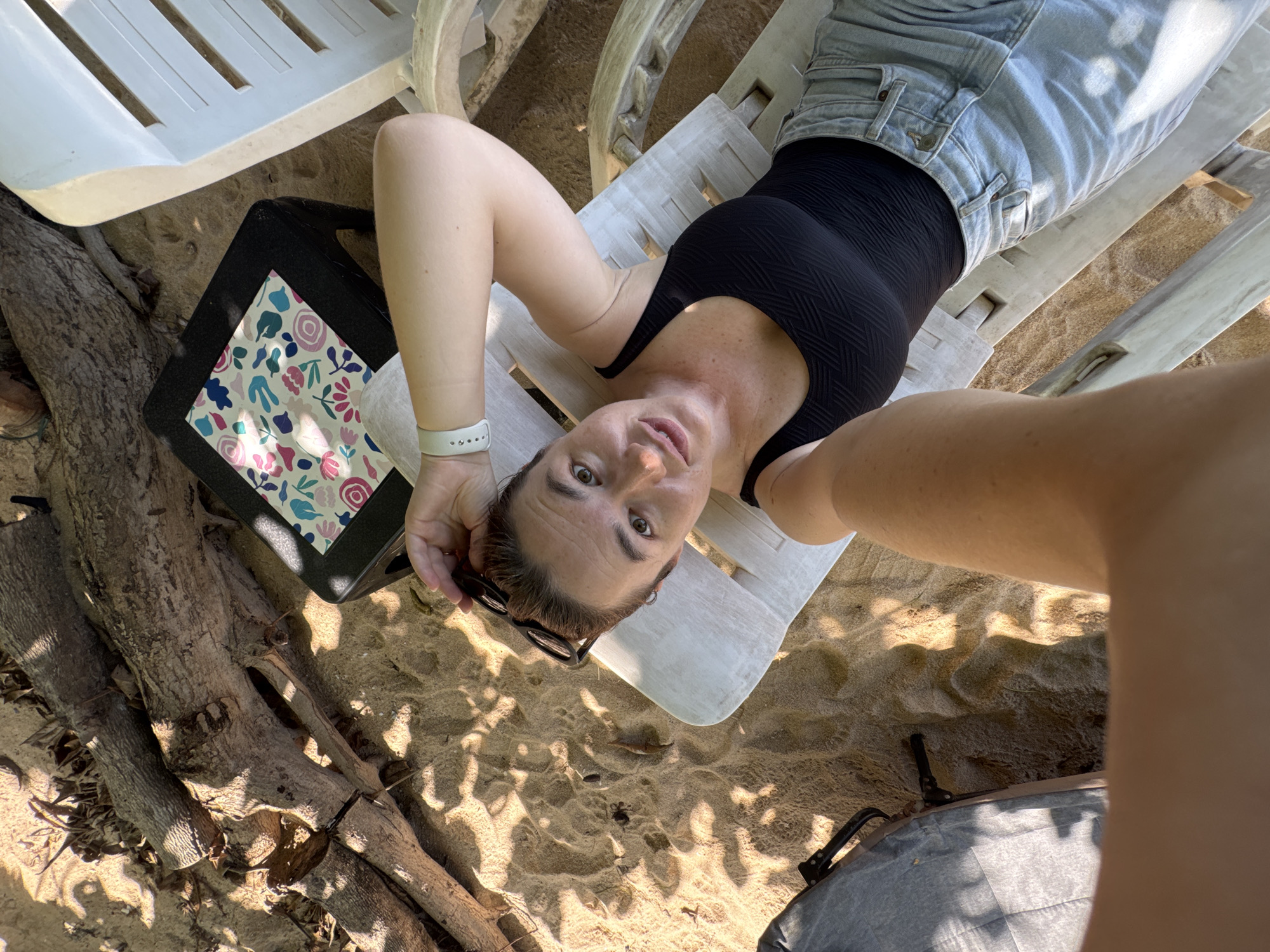 A woman is lying on a white beach chair on a sandy beach. She is wearing a black swimsuit and light blue denim shorts. The woman is holding sunglasses in her right hand and resting her head on her arm. Her left arm is extended towards the camera, with a white fitness tracker on her wrist. Behind her, there is a small black table with a patterned cloth on top. Several pieces of driftwood are scattered on the sand. The scene is illuminated by sunlight, casting shadows on the sand and surrounding objects. The location appears to be a beach in Rawai, Thailand.