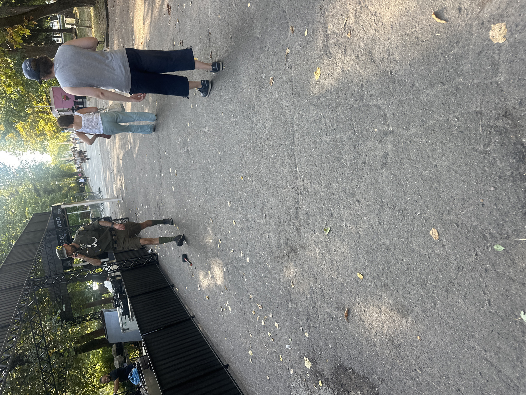 The image captures a scene on a paved walkway lined with trees in the city of Ternopil, Ukraine. In the foreground, a man in a military-style uniform with shorts and knee-high socks is walking and speaking on a cell phone. A black object resembling a hammer lies on the ground near his feet. Further down the walkway, two individuals are walking away from the camera: a woman in a white top and light blue pants, and a man in a grey sleeveless shirt and dark shorts. The background shows a crowd of people, tents, and trees, suggesting an outdoor event or festival. The lighting indicates it is daytime, possibly late afternoon given the long shadows.