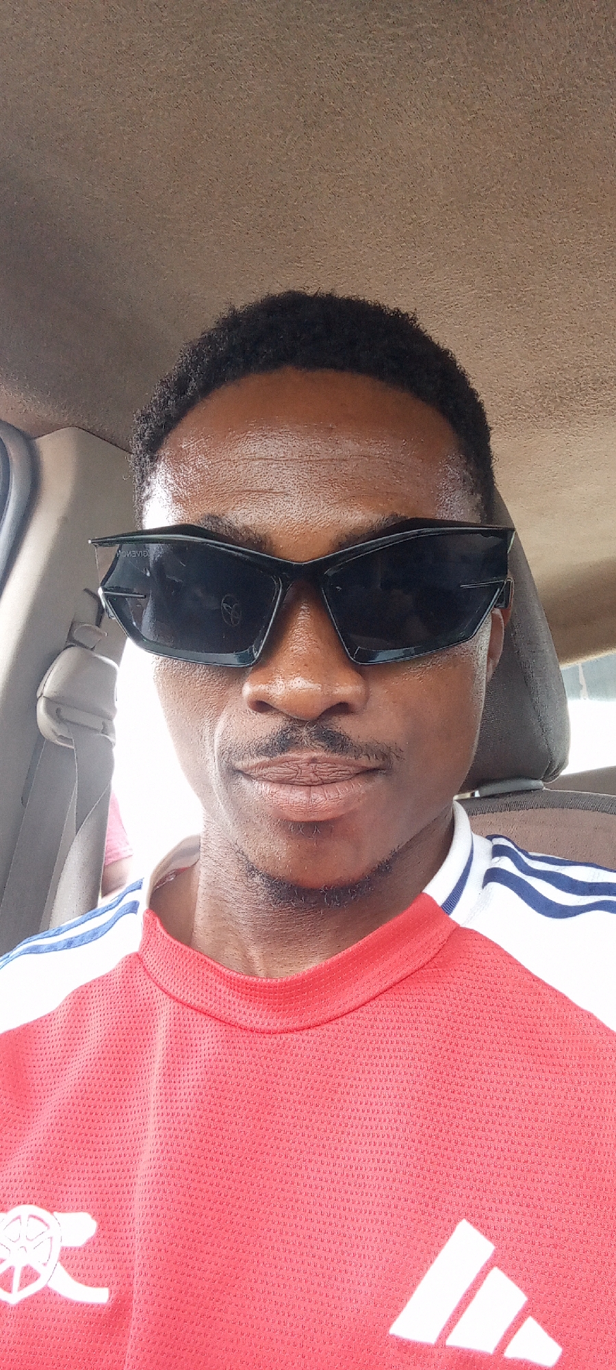 A medium, front-view shot of a man sitting in a car in Otunade, Nigeria. He is wearing black sunglasses, a red Arsenal football jersey with white stripes on the shoulders, and has a neatly trimmed mustache and beard. The interior of the car is visible, including the seat belt and car seat headrest. The background shows the car's interior.