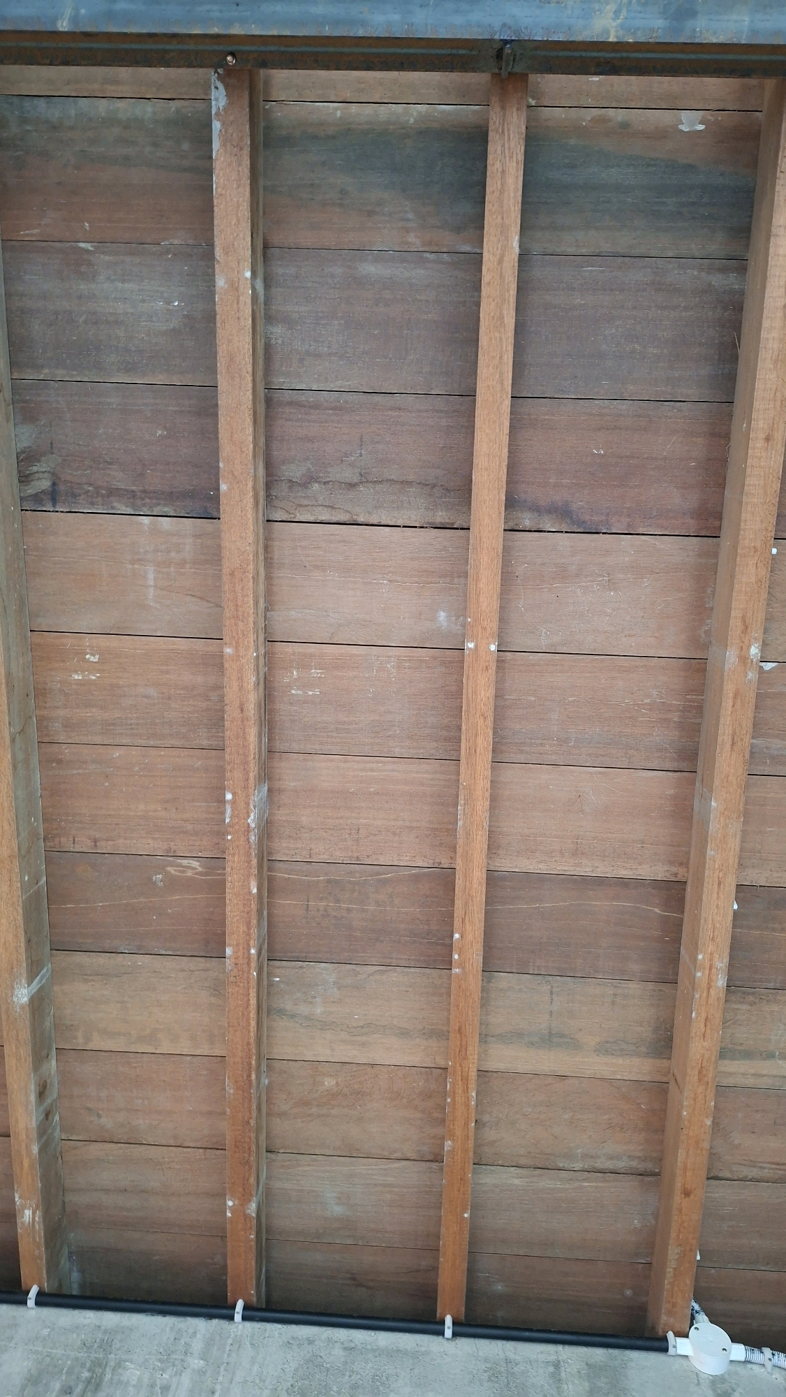 The image shows a close-up, eye-level view of a wooden ceiling structure. The ceiling is made of horizontal wooden planks, stained a reddish-brown, with visible grain and some wear. Running vertically across the planks are three wooden support beams. A dark metal beam is visible at the top of the image, spanning horizontally. At the bottom of the image, a black pipe, supported by white clips, runs horizontally along what appears to be a concrete surface. The scene appears to be an outdoor or semi-outdoor structure, possibly a roof or a pergola. There are no people visible in the image. The location is described as Gronggrong, Indonesia.
