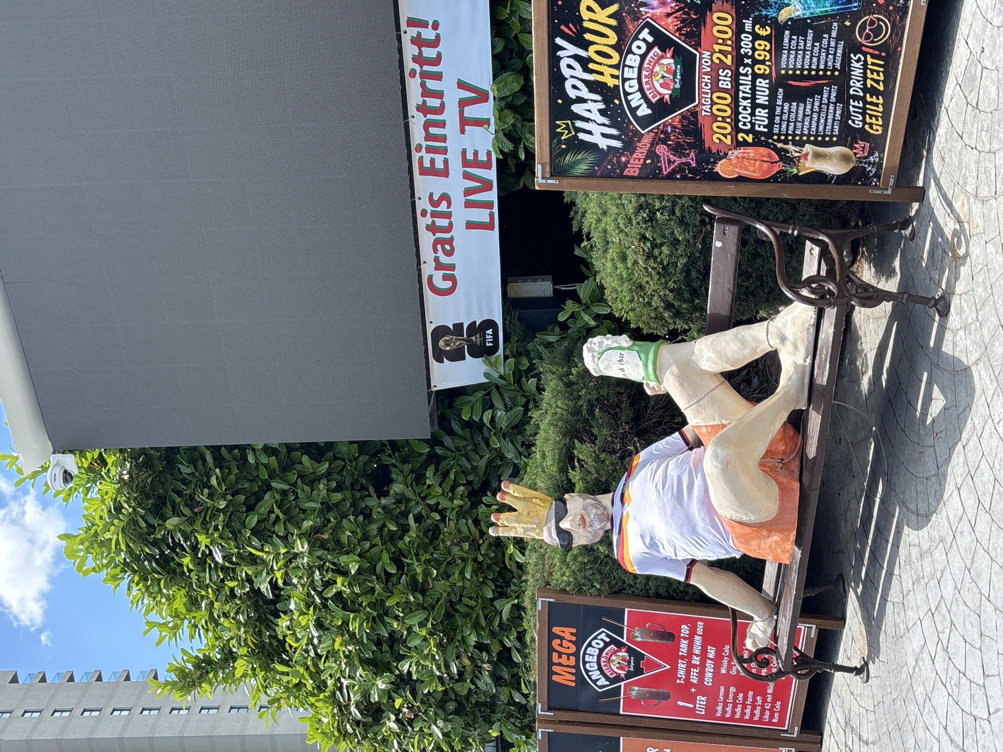 The image depicts an outdoor scene in Varna, Bulgaria, featuring a large television screen mounted above a row of promotional boards and a mannequin. The mannequin, dressed in a white shirt with red and black trim, orange shorts, sunglasses, and a gold crown, is seated on a brown metal bench. To the left of the mannequin is a promotional board with the text "MEGA ANGEBOT" and "BIERKONIG" along with images of drinks and a list of offerings including "T-SHIRT, TANK TOP, APFE, BK HUHN ODER COWBOY HAT" and various alcoholic drinks. To the right of the mannequin is another promotional board that reads "HAPPY HOUR ANGEBOT BIERKONIG" and lists "2 COCKTAILS x 300 ml. FÜR NUR 9,99 €" along with a menu of cocktails and the slogan "GUTE DRINKS GEILE ZEIT!". Behind the mannequin and boards is lush green foliage. In the background, a tall, modern building with multiple windows and balconies is visible, as is a blue sky with scattered clouds. A white security camera is mounted on the exterior wall of the building in the upper left portion of the image. The ground is paved with circular patterned stones.