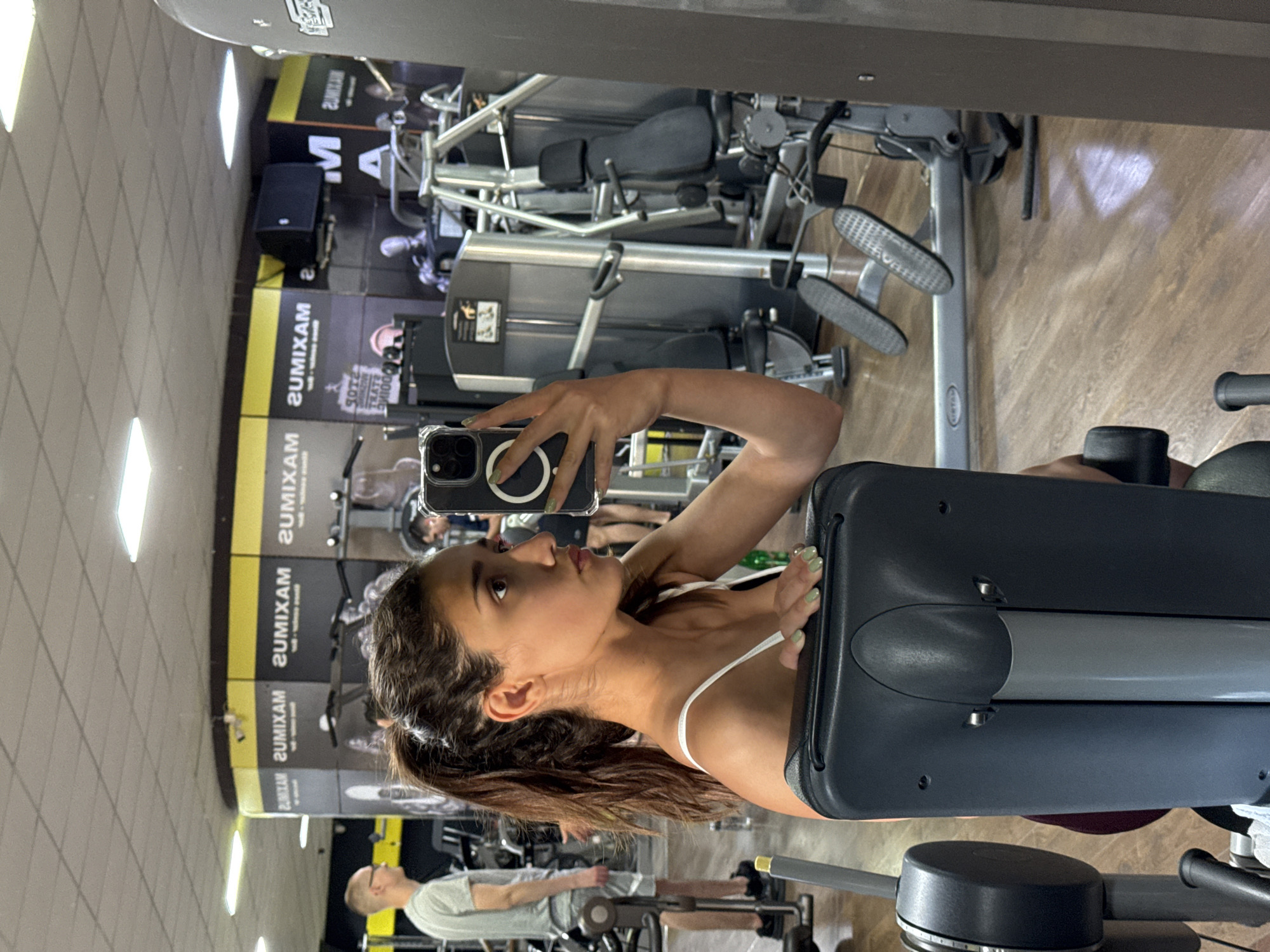 A young woman is taking a selfie in a gym. The gym appears to have a variety of weight machines. In the background, a man is exercising on a machine. "MAXIMUS" is visible on the wall, potentially the name of the gym. The gym is located in Bar, Montenegro.