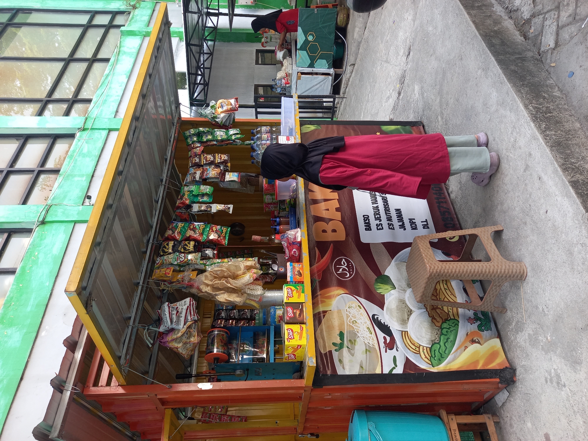 In Ploso, Indonesia, a red and yellow food stall is set up on a concrete sidewalk. The stall, likely a converted shipping container, displays various snack bags and boxed drinks hanging from its awning. A blue plastic bin is situated to the left of the stall. In front of the stall, a young person wearing a red tunic and light green pants stands facing away from the camera. A brown plastic stool is placed near the front of the stall. The stall has a large banner with an illustration of a bowl of noodles and meatballs, along with text in Indonesian. To the right of the food stall, another stall is partially visible with a woman in a red hijab serving a customer. The background shows a building with green painted walls and large windows.