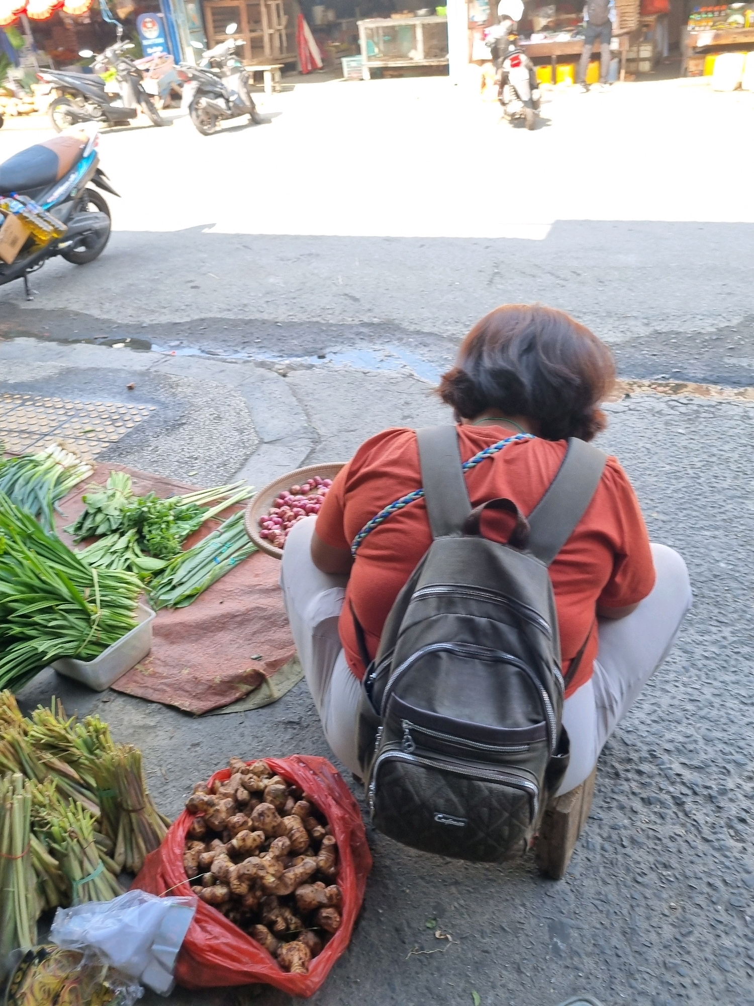 A woman with dark, short hair is squatting on a wooden stool on an asphalt surface. She is facing away from the camera and wearing an orange shirt and light-colored pants. A dark green backpack is on her back. In front of her, on the ground, is a red plastic bag filled with ginger root. To her left, there are bundles of green vegetables, some in a white plastic container and others tied with rubber bands. A woven basket containing small red onions is also visible near her. In the background, there are several motorcycles parked and market stalls, suggesting a street market environment in Tatura Utara, Indonesia.
