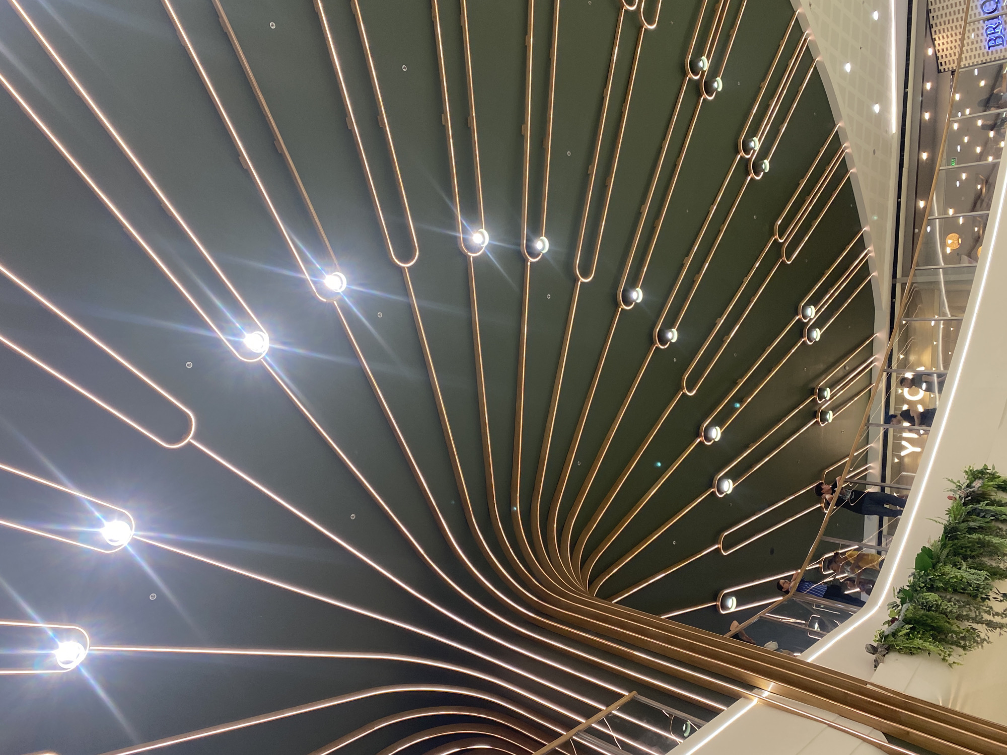 The image shows an interior view of a shopping mall in Davao City, Philippines, specifically in the Abreeza Ayala Malls. The ceiling features a distinctive design with numerous parallel, curved copper-colored tubes illuminated by embedded lights. These tubes radiate outwards from a central point, creating a dynamic, fan-like pattern against a dark green background. Several individuals are visible on a mezzanine level, leaning on a glass railing. One person, wearing a blue and black striped shirt, stands near the railing, while another, in a yellow garment, is seated. A person in a black shirt is also present. The environment includes lush green artificial plants cascading down from the upper level, and a white staircase with a glass balustrade. The overall impression is of a modern and architecturally interesting retail space.