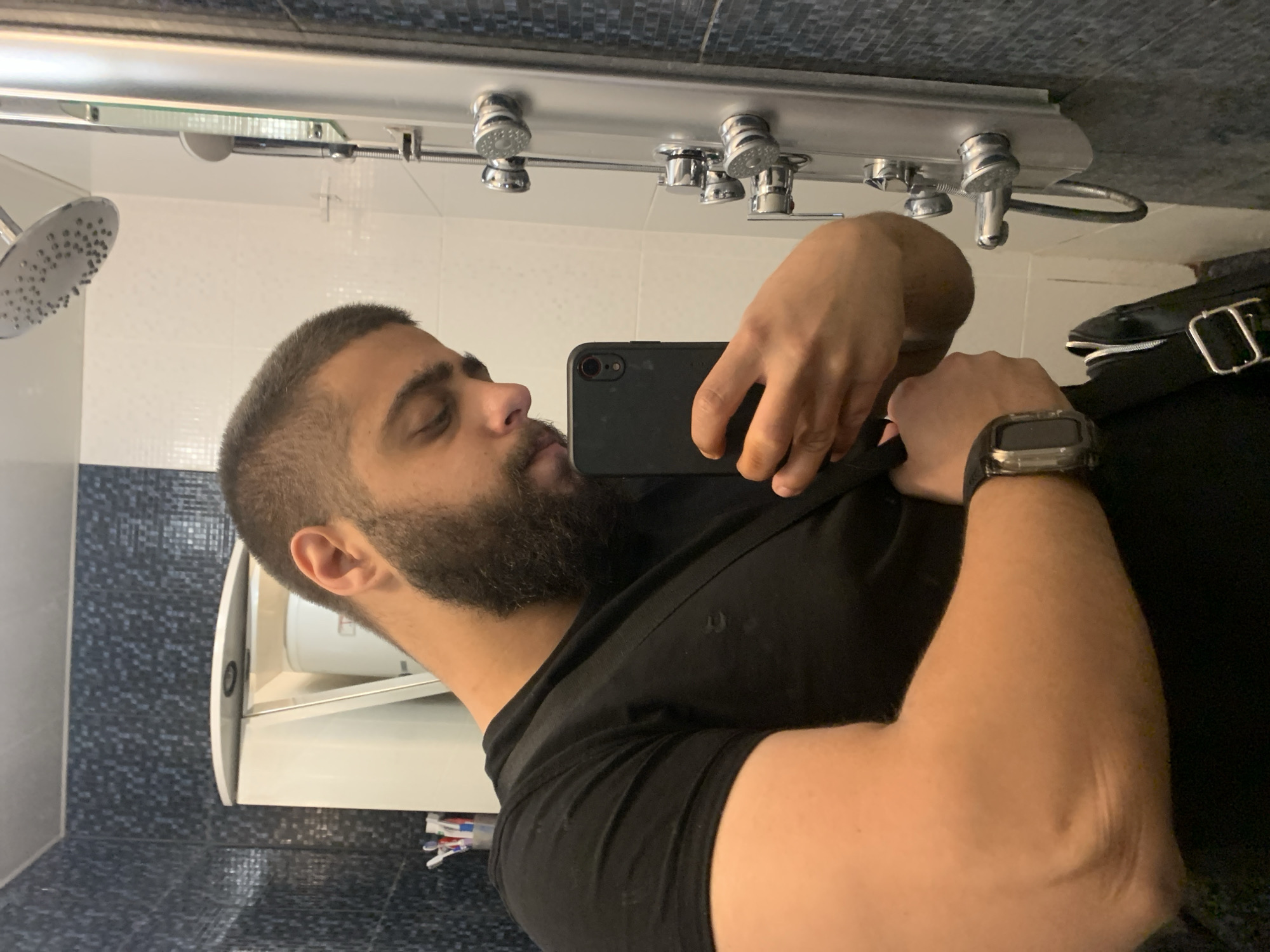 A mirror selfie captures a man in a modern bathroom setting. The man has a short beard and dark hair with a faded haircut. He is wearing a black t-shirt and a dark digital watch on his left wrist. He holds a dark-colored smartphone, with its camera visible, to take the picture, looking towards his reflection.
The background reveals typical bathroom features. To the man's left, a silver rain shower head and multiple chrome body jets are mounted on a metallic panel. The walls are tiled, featuring a section of small, dark blue/black mosaic tiles at the top and bottom, with larger white tiles in between. To the man's right, a white wall-mounted sink or vanity unit with an open cabinet door is visible, and part of a white toilet bowl can be seen behind him. A black bag or strap with a silver buckle is partially visible on the far right.
The activity is the man taking a mirror selfie. The setting appears to be a well-lit indoor environment, possibly a hotel, gym, or contemporary home bathroom, given the style of the fixtures. There is no visible text in the image.