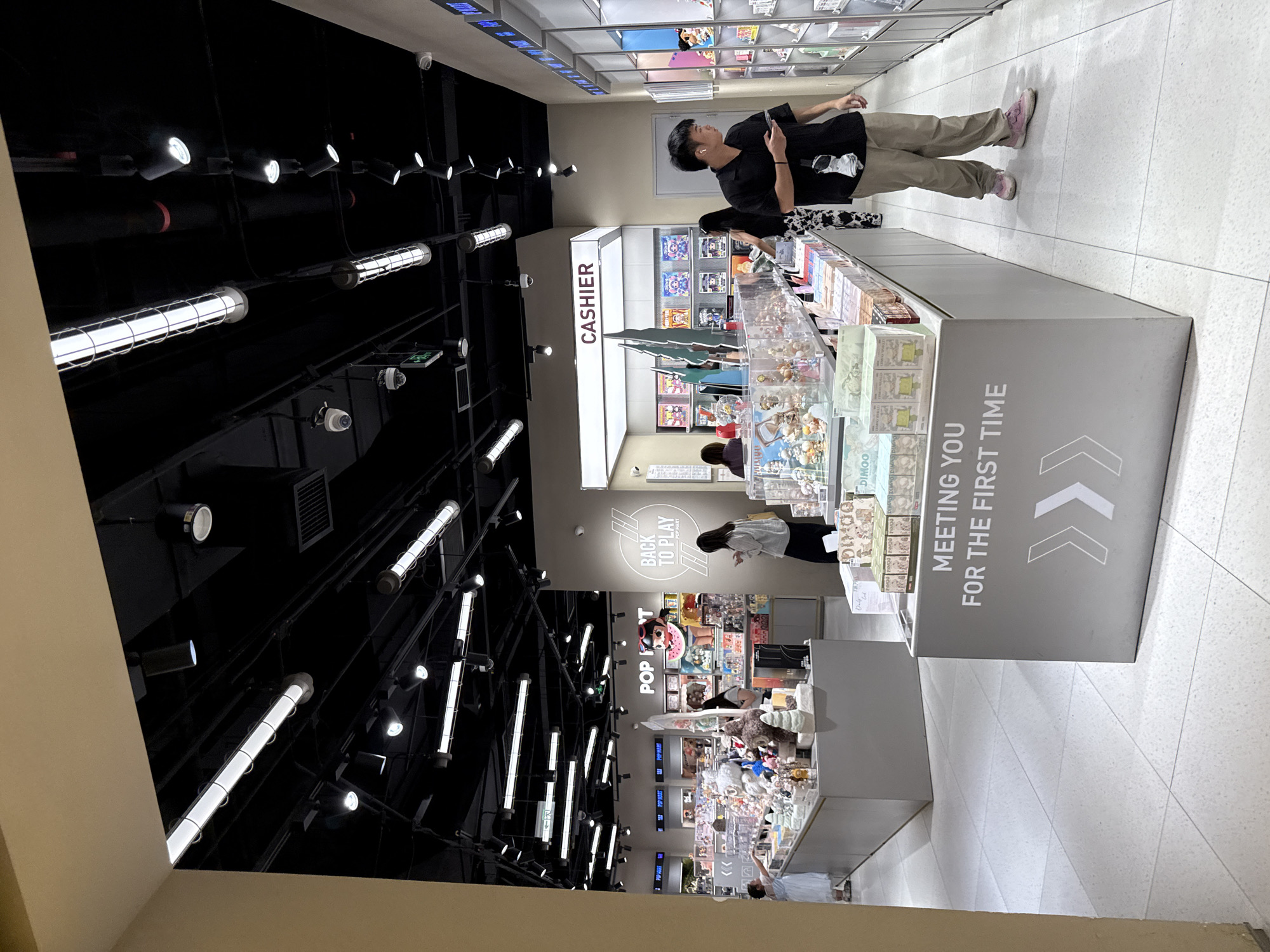 The image depicts the interior of a retail store, likely a "Pop Mart" store based on signage. The store appears to be a modern, well-lit space with a black ceiling featuring numerous track lights and tube lights. The flooring is tiled in a light color. In the foreground, there is a grey display counter with the text "MEETING YOU FOR THE FIRST TIME" and arrow graphics.
Several people are visible within the store. A young man wearing a black shirt and tan pants is looking at his phone while walking. Another person, possibly a woman, is standing to the right, looking into display cases. Further into the store, a woman in a white top and black skirt is standing near a display, and another woman in a white dress is browsing merchandise on the left.
The store is filled with display shelves and counters showcasing a variety of items, likely collectible toys or figurines, judging by the colorful packaging and the nature of the merchandise visible. There are also signs indicating "CASHIER" and "BACK TO PLAY POP MART". The overall impression is of a bustling retail environment focused on popular collectible merchandise. The location is identified as Haizhu District, China.