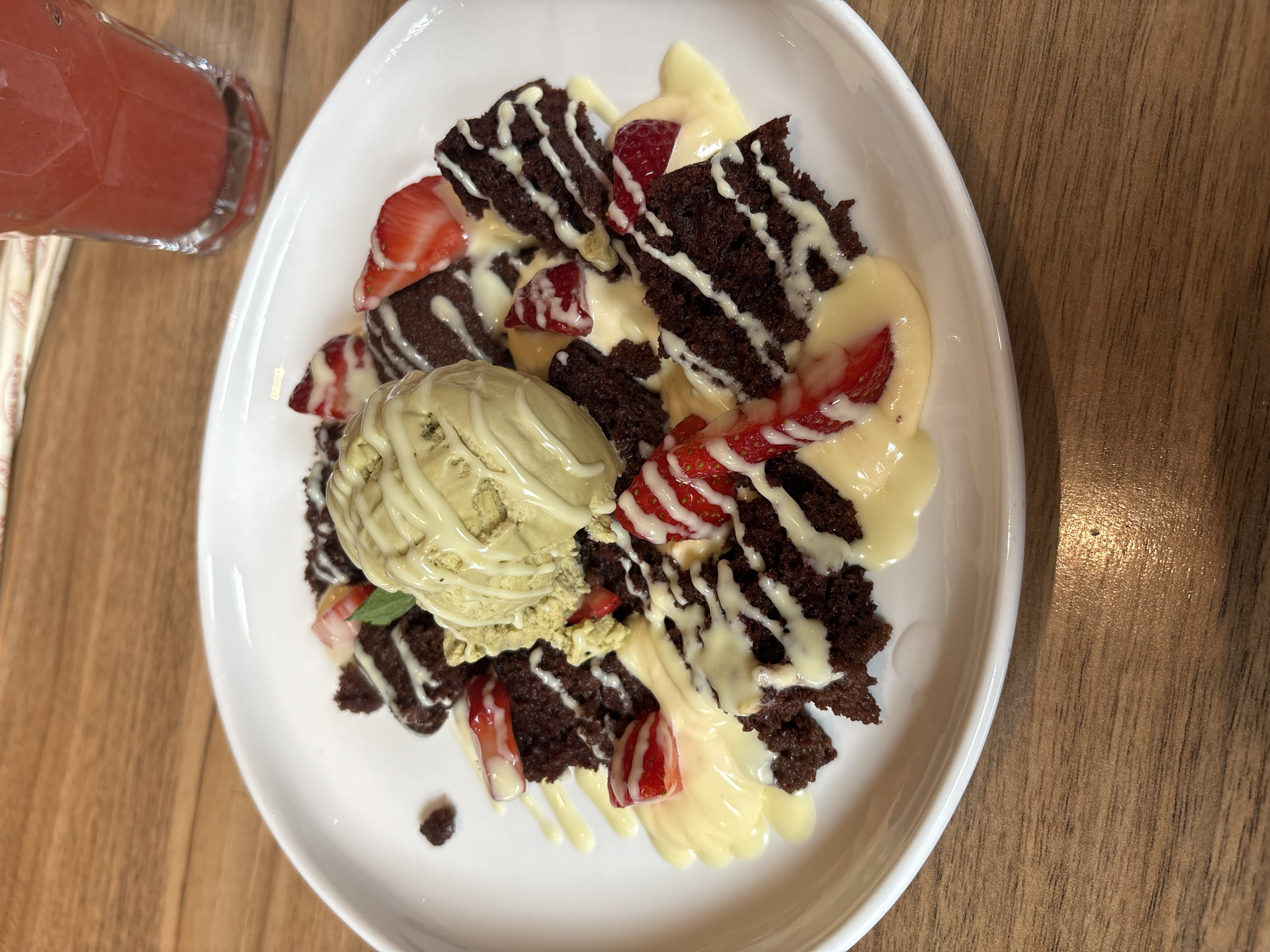 A top-down view depicts a dessert on a white plate, placed on a wooden table. The dessert consists of brownie pieces drizzled with white icing and vanilla sauce. Sliced strawberries are scattered throughout. A scoop of green ice cream is centrally positioned. A glass of pink drink and chopsticks are visible on the left side of the plate. The location is unspecified.