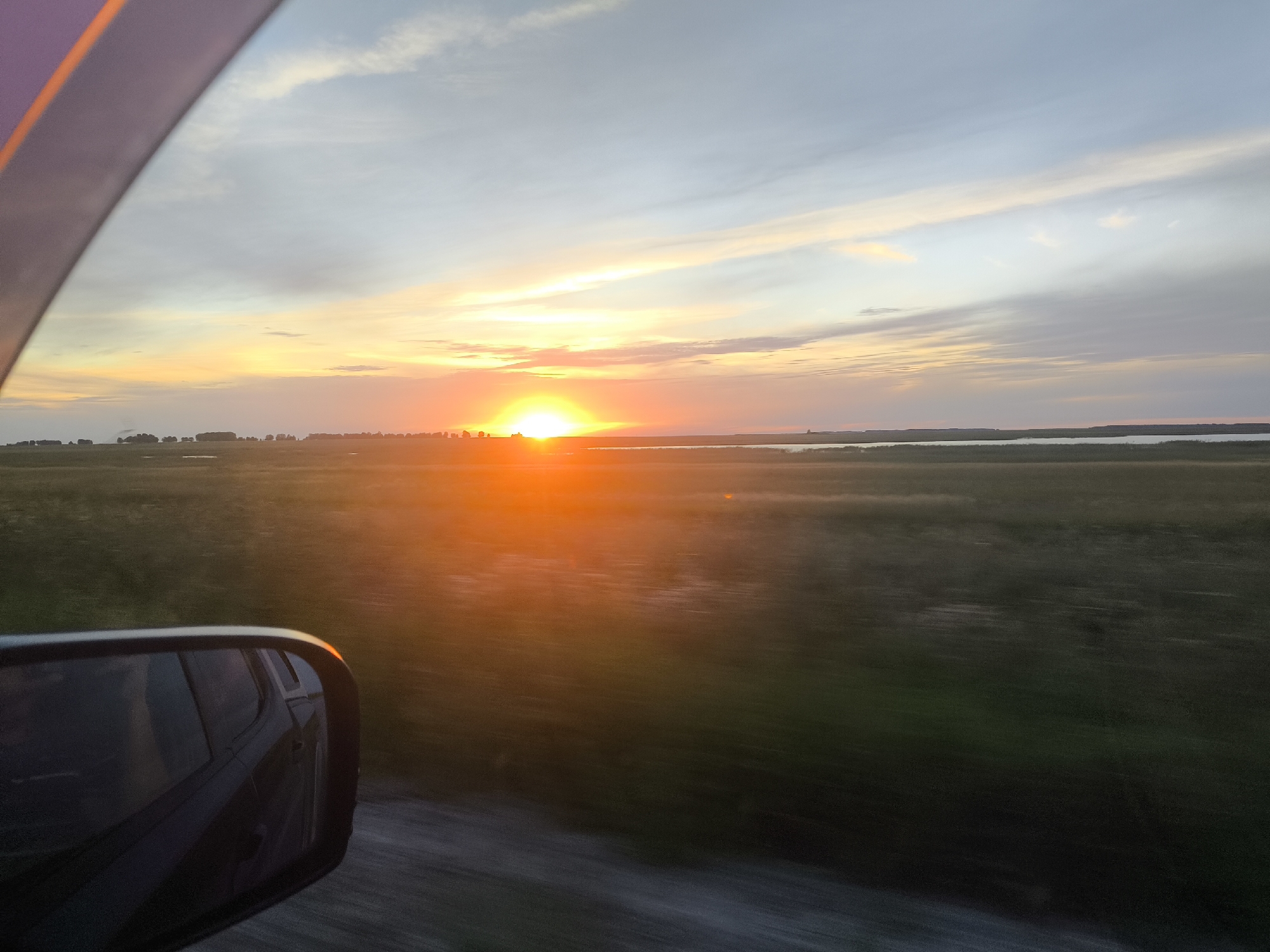 The image captures a sunset over a rural landscape, viewed from the interior of a moving vehicle. The setting sun, a bright orange orb, is low on the horizon, casting a warm glow across the sky. The sky transitions from a pale blue at the top to shades of orange, yellow, and pink near the sun, with wispy clouds scattered throughout.

The foreground features the side mirror of a vehicle, reflecting the car's interior and the surrounding scenery in a distorted manner due to motion. The mirror is mounted on the car's body, which is partially visible on the left side of the frame.

Beyond the vehicle, the landscape is characterized by flat, grassy fields with sparse trees dotting the distant horizon. A body of water, possibly a lake or river, is visible in the middle ground, reflecting the sunset colors. The motion blur suggests the image was taken while driving on a dirt or gravel road, indicated by the textured surface in the lower part of the frame.

The scene is set in the Пресноредуть ауылдық округі, Kazakhstan, a region known for its vast plains and agricultural lands, which aligns with the visual elements present in the photograph. There are no people or specific actions clearly depicted, aside from the implied movement of the vehicle.