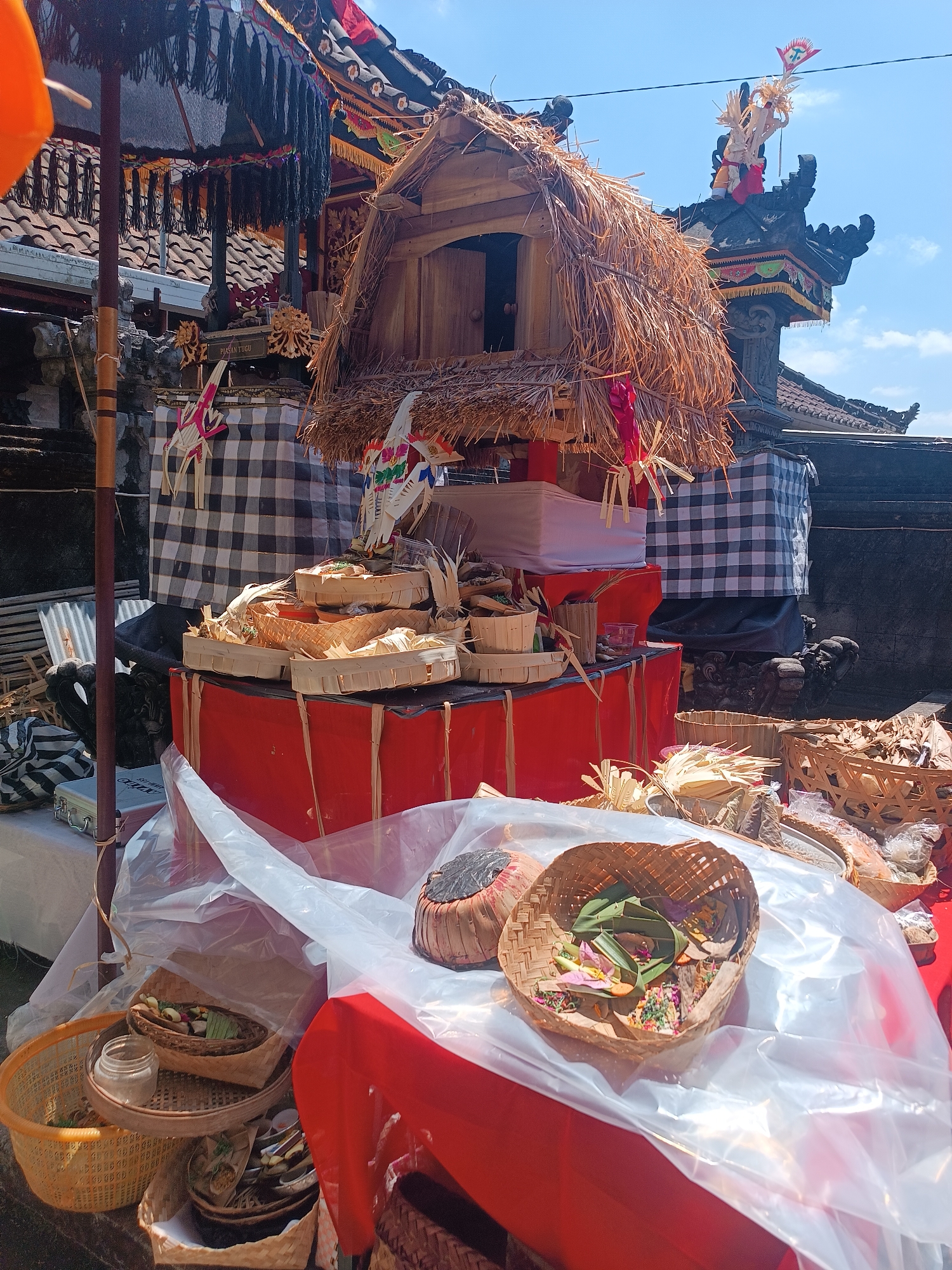 The image depicts an outdoor market stall in Kerambitan, Indonesia, likely during a cultural or religious event, given the offerings displayed. The stall is adorned with traditional Balinese decorations, including black and white checkered fabric, woven baskets filled with offerings, and a tiered structure topped with a miniature thatched-roof building. These offerings appear to be made of leaves, flowers, and possibly food items, arranged in small woven containers. A large, dark umbrella with fringe hangs in the foreground, partially obscuring a stack of red boxes. Behind the stall, a traditional Balinese temple structure is visible with ornate carvings and a tiled roof. The ground is a mix of dark earth and concrete. The scene is illuminated by bright sunlight, casting shadows and highlighting the vibrant colors of the offerings and decorations. No people are clearly visible in the immediate foreground or center of the image.